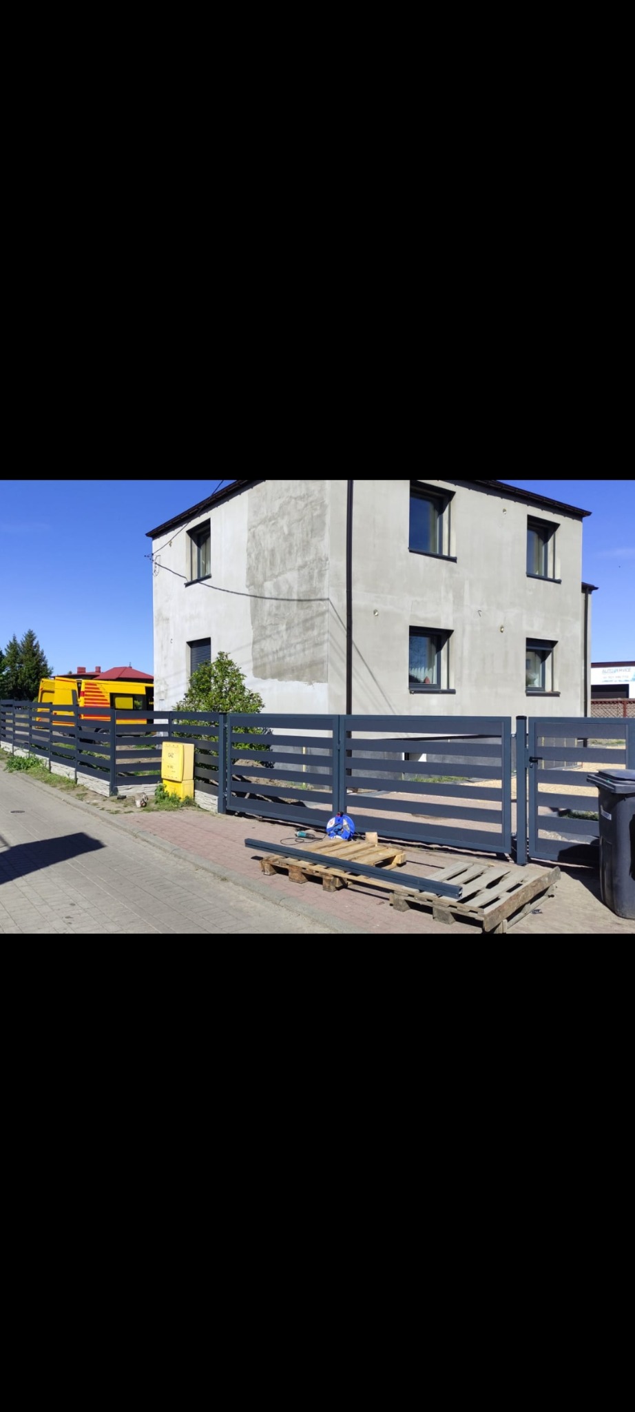 Nowoczesne, poziome ogrodzenie metalowe w kolorze grafitowym przed domem z szarą elewacją. Na pierwszym planie paleta i narzędzia, sugerujące świeży montaż.