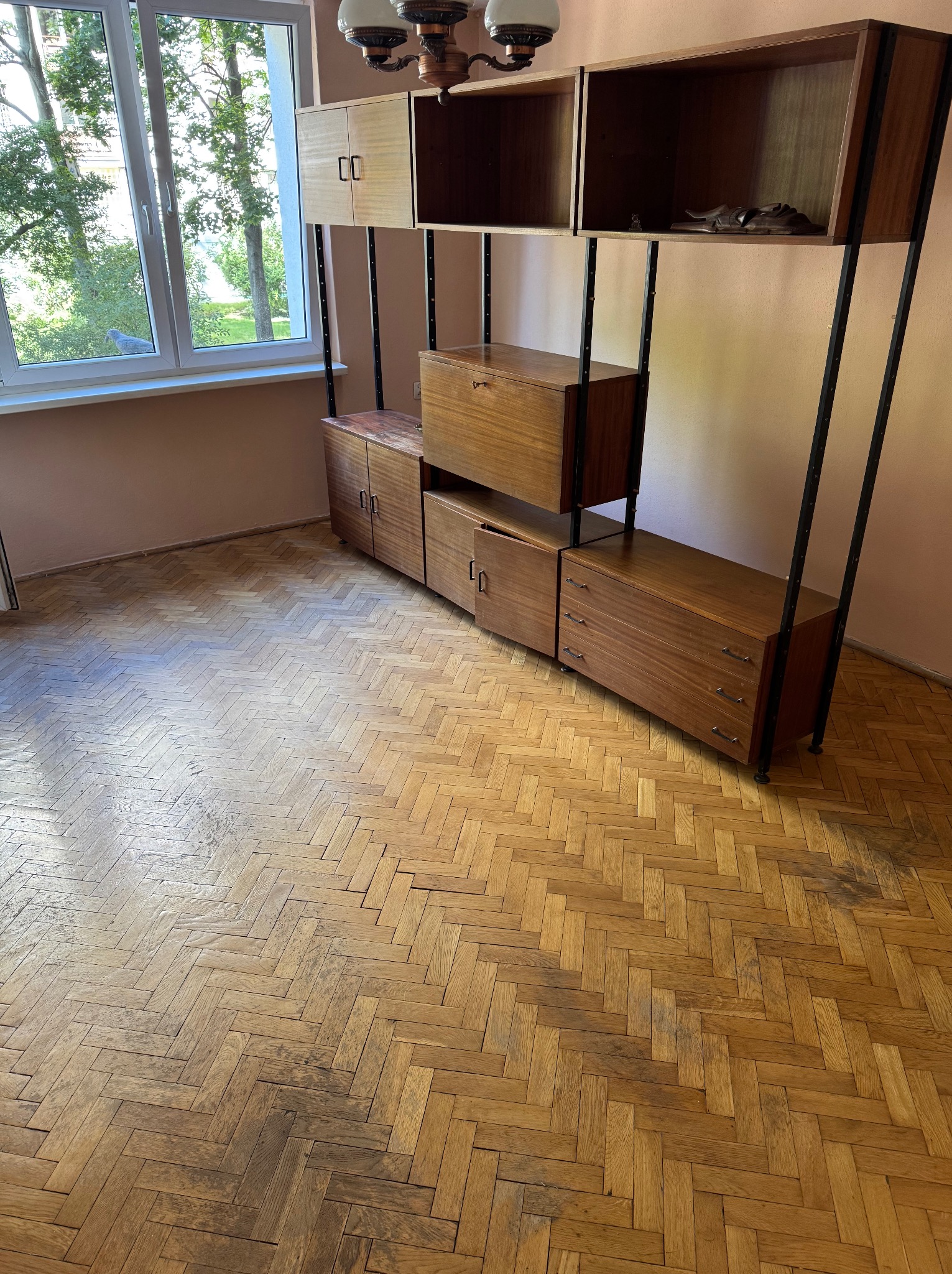 Parkiet ułożony w jodełkę po renowacji, widoczny fragment regału w stylu vintage, okno z widokiem na zieleń. Jasne, naturalne światło podkreśla odnowioną powierzchnię podłogi.