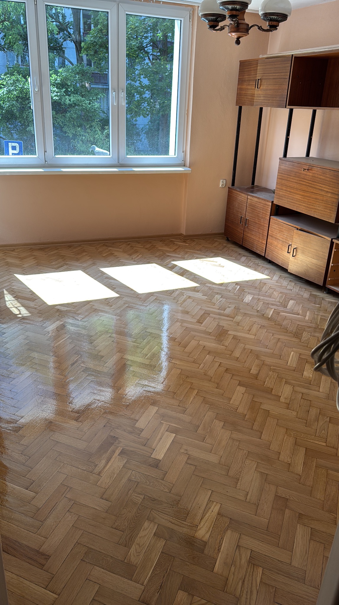 Parkiet ułożony w jodełkę po renowacji, z widocznymi refleksami światła słonecznego. W tle okno i meble w stylu vintage. Jasne, naturalne wykończenie drewna.