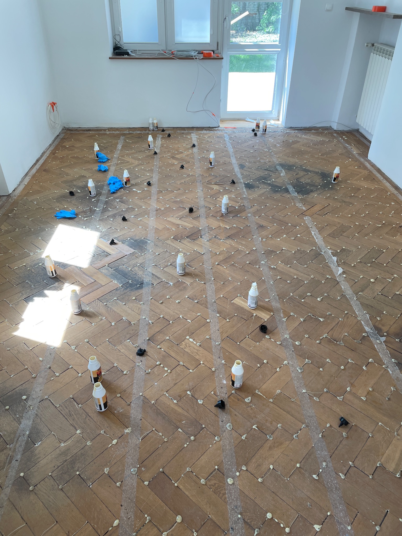 Parkiet w jodełkę po naprawie, z widocznymi śladami kleju i buteleczkami preparatu. Przygotowanie do dalszej obróbki, jasne wnętrze z oknem.