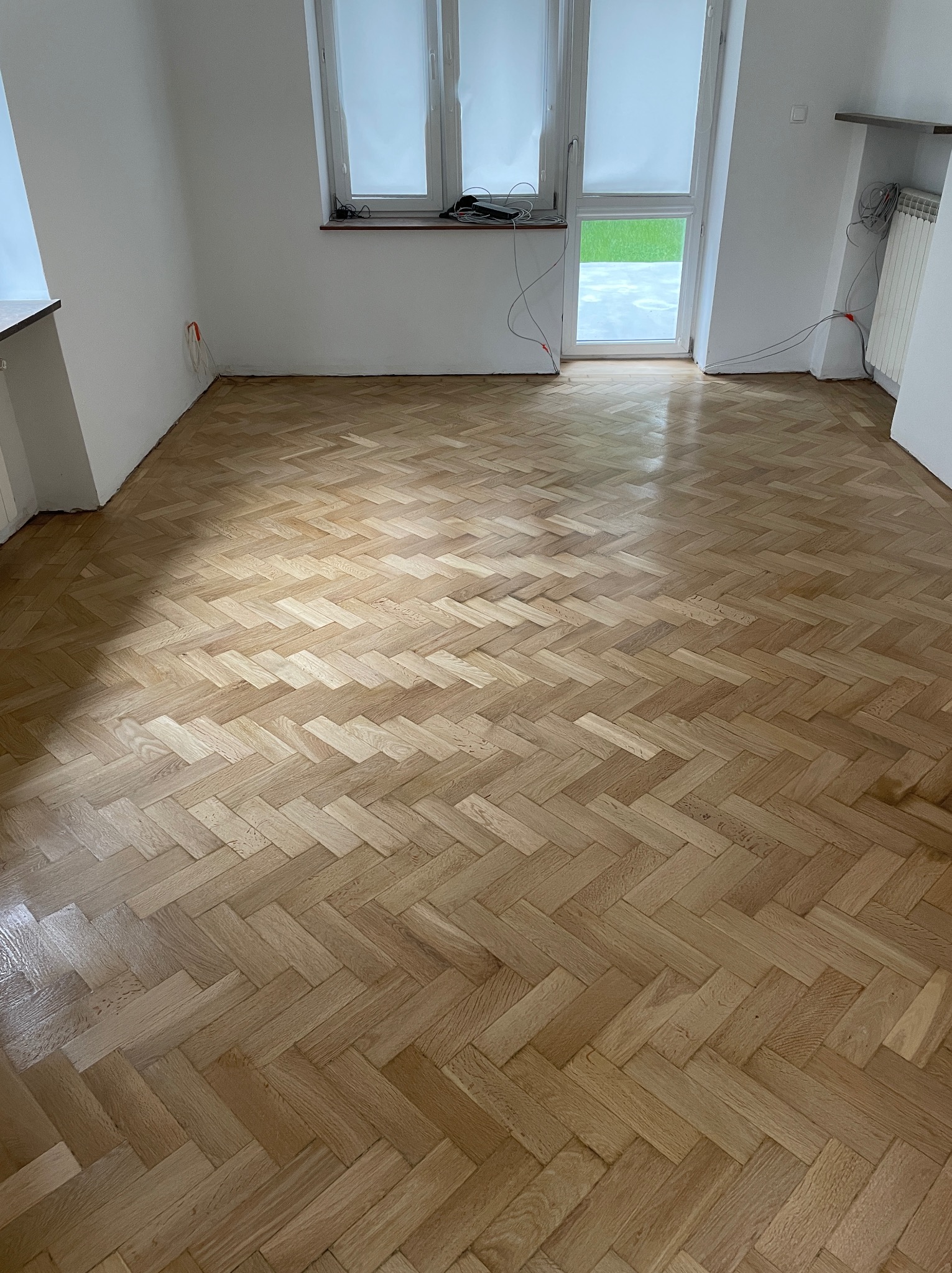 Jasny parkiet ułożony w jodełkę po renowacji w pustym pokoju z białymi ścianami i oknem. Widoczne ślady po cyklinowaniu, podkreślone przez padające światło.