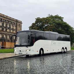 V8XD SPÓŁKA Z OGRANICZONĄ ODPOWIEDZIALNOŚCIĄ - Biały autokar Mercedes-Benz V8XD na mokrym bruku, z budynkiem w tle. Widoczne rowery miejskie i osoby z parasolami. Pochmurny dzień.