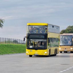 V8XD SPÓŁKA Z OGRANICZONĄ ODPOWIEDZIALNOŚCIĄ - Żółty, dwupiętrowy autobus MAN z pasażerami, jadący po betonowym placu. W tle drugi autobus Mercedes w kolorze złotym. Widoczne drzewa i niebo.