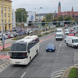 V8XD SPÓŁKA Z OGRANICZONĄ ODPOWIEDZIALNOŚCIĄ - Biały autokar na tle miejskiego krajobrazu z wieżą kościoła. Ulica z ruchem samochodowym, widoczne budynki i drzewa. Dzień, pochmurne niebo.