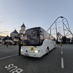 V8XD SPÓŁKA Z OGRANICZONĄ ODPOWIEDZIALNOŚCIĄ - Biały autokar Mercedes-Benz V8XD na parkingu z napisem 'BUS', w tle park rozrywki z kolejką górską i zamkiem. Tabliczka z symbolem dzieci.