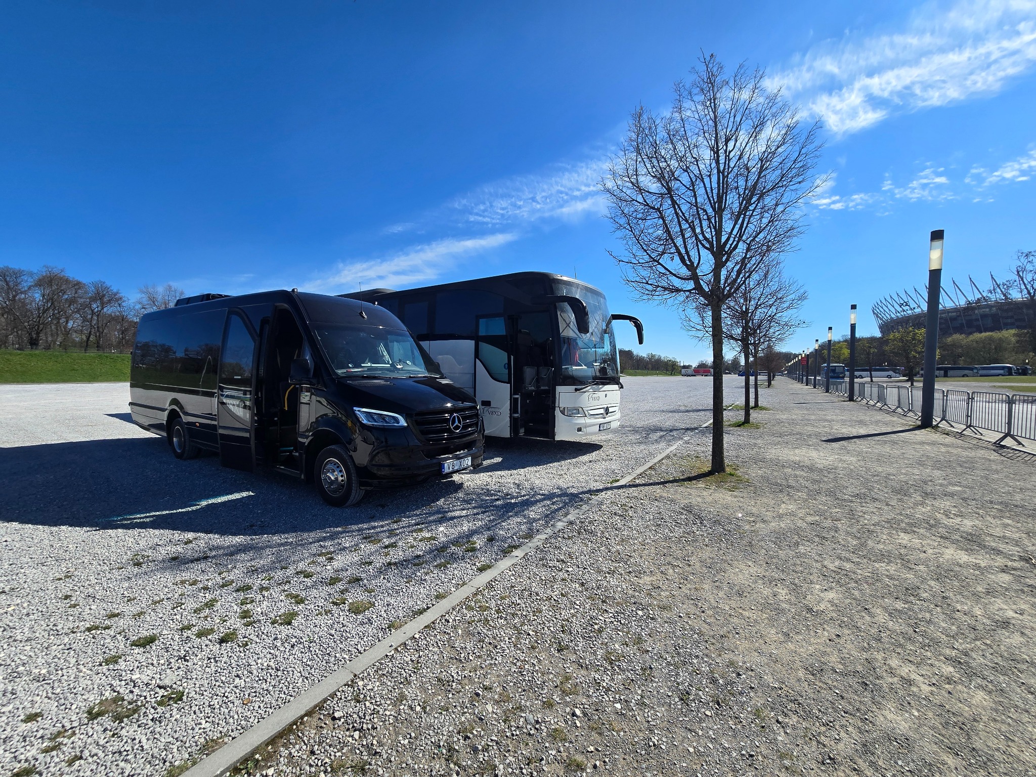 Czarny bus Mercedes z otwartymi drzwiami i biały autokar na żwirowym parkingu, błękitne niebo z chmurami w tle. Stadion Narodowy w oddali.