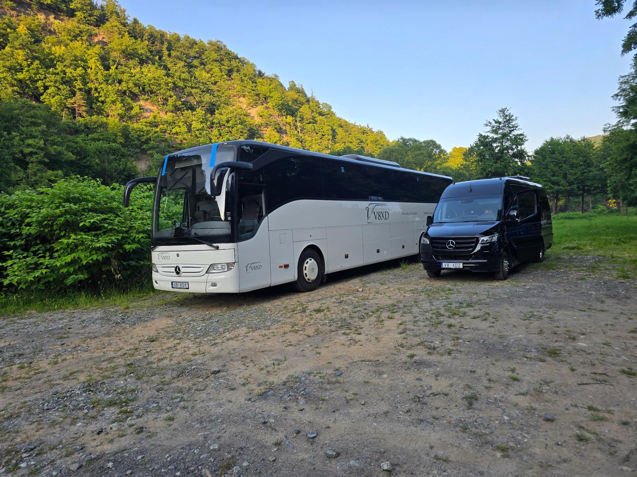 Autokar i bus Mercedes-Benz V8XD na żwirowym podłożu, w tle zalesione wzgórze. Autokar biały z czarnymi akcentami, bus czarny. Dzień, naturalne światło.