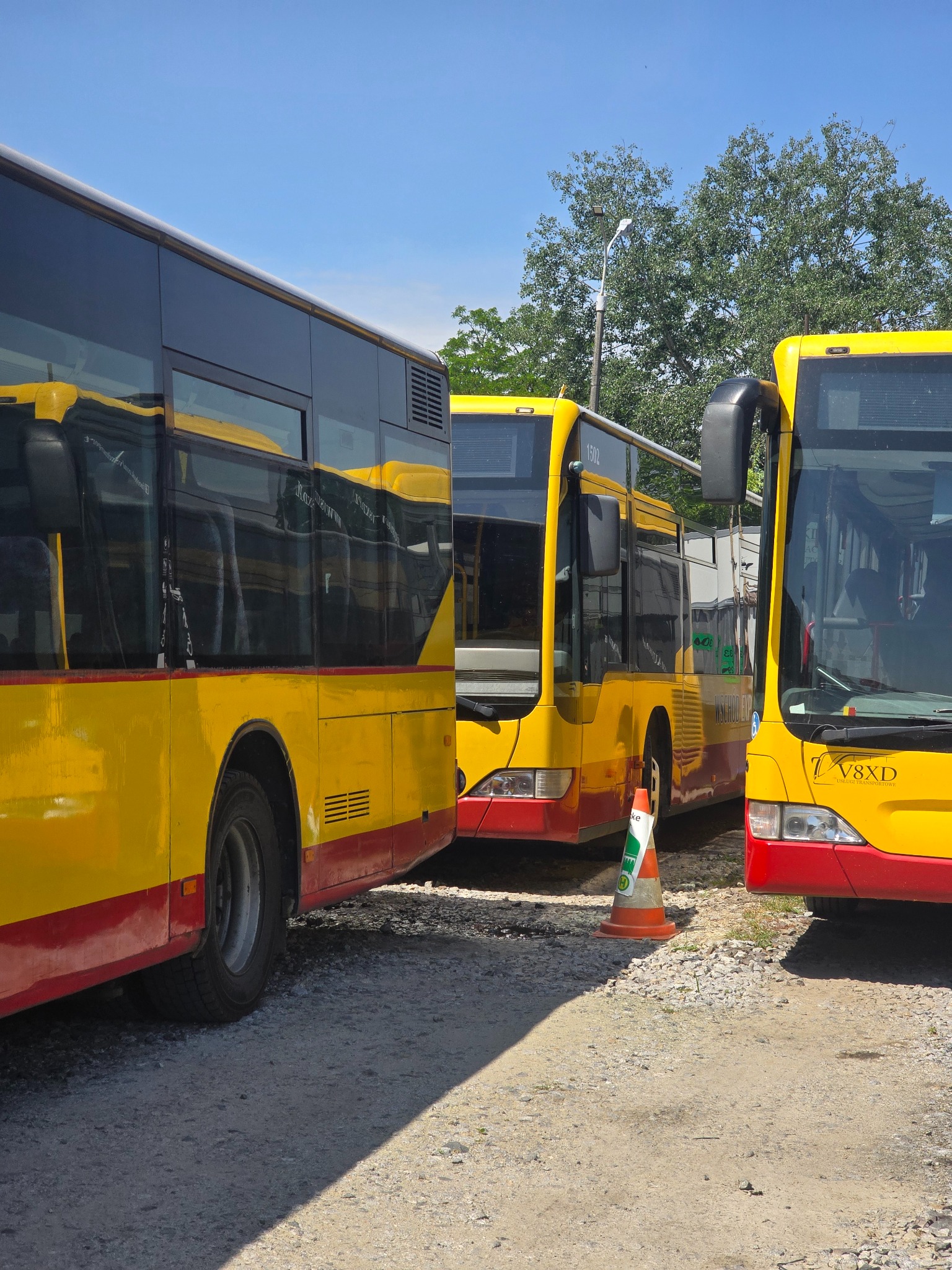 Żółto-czerwone autobusy miejskie stojące na żwirowym placu w słoneczny dzień, z widocznym stożkiem ostrzegawczym i drzewami w tle. Autobusy z numerami linii.
