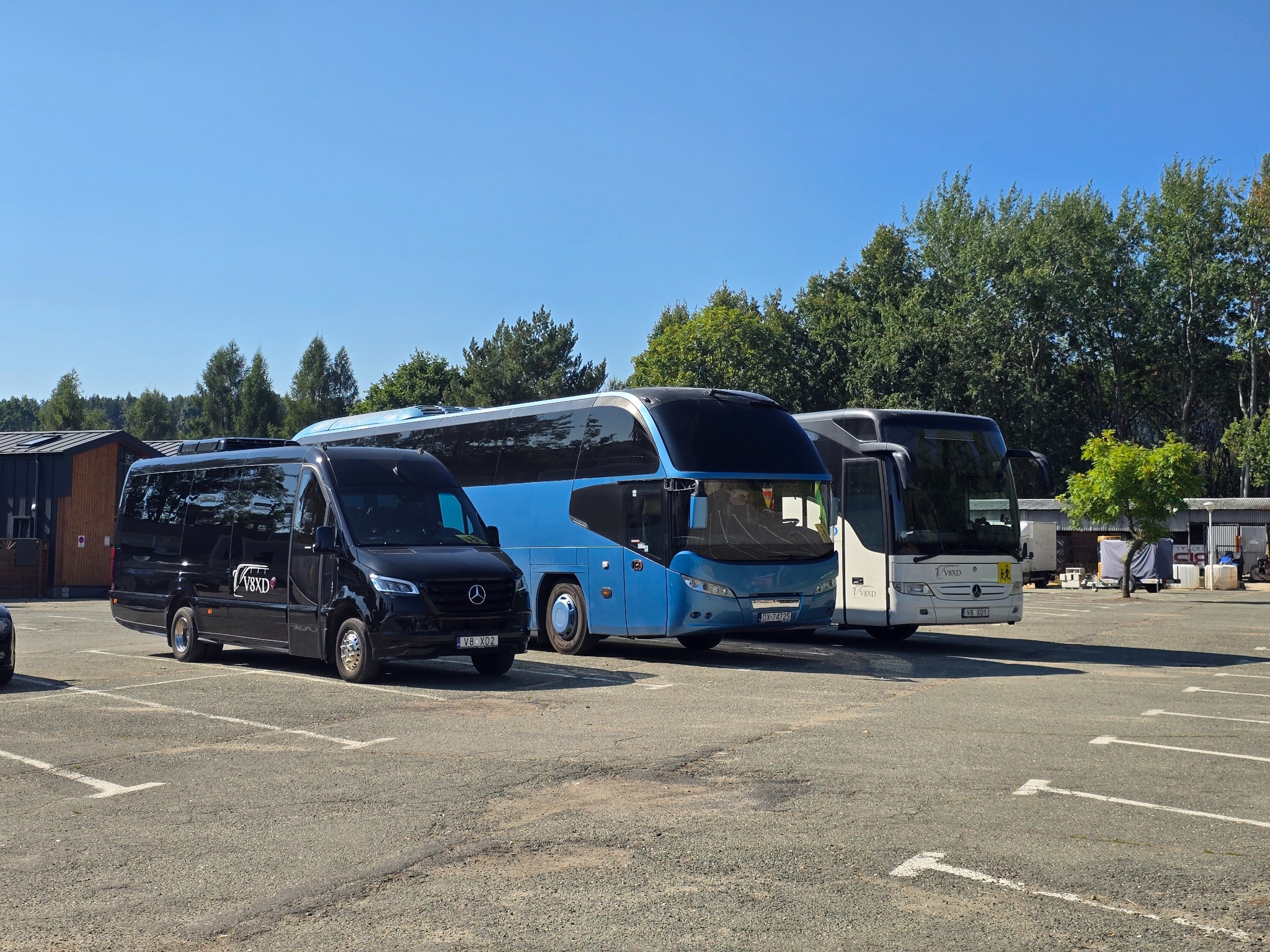 Flota pojazdów firmy transportowej: czarny bus Mercedes, niebieski autokar i biały autokar na parkingu w słoneczny dzień. Widoczne logo firmy V8XD.