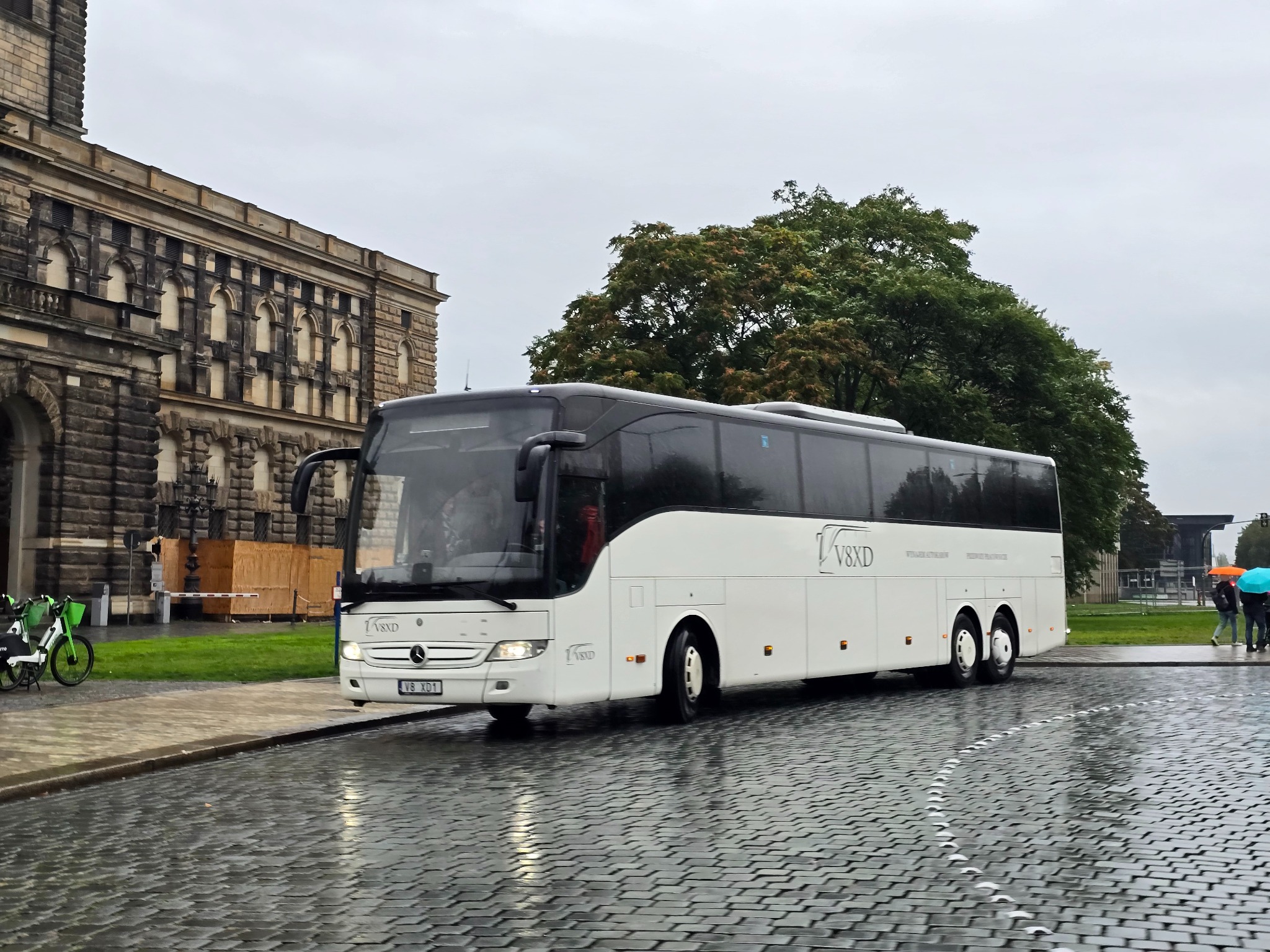 Biały autokar Mercedes-Benz V8XD na mokrym bruku, z budynkiem w tle. Widoczne rowery miejskie i osoby z parasolami. Pochmurny dzień.