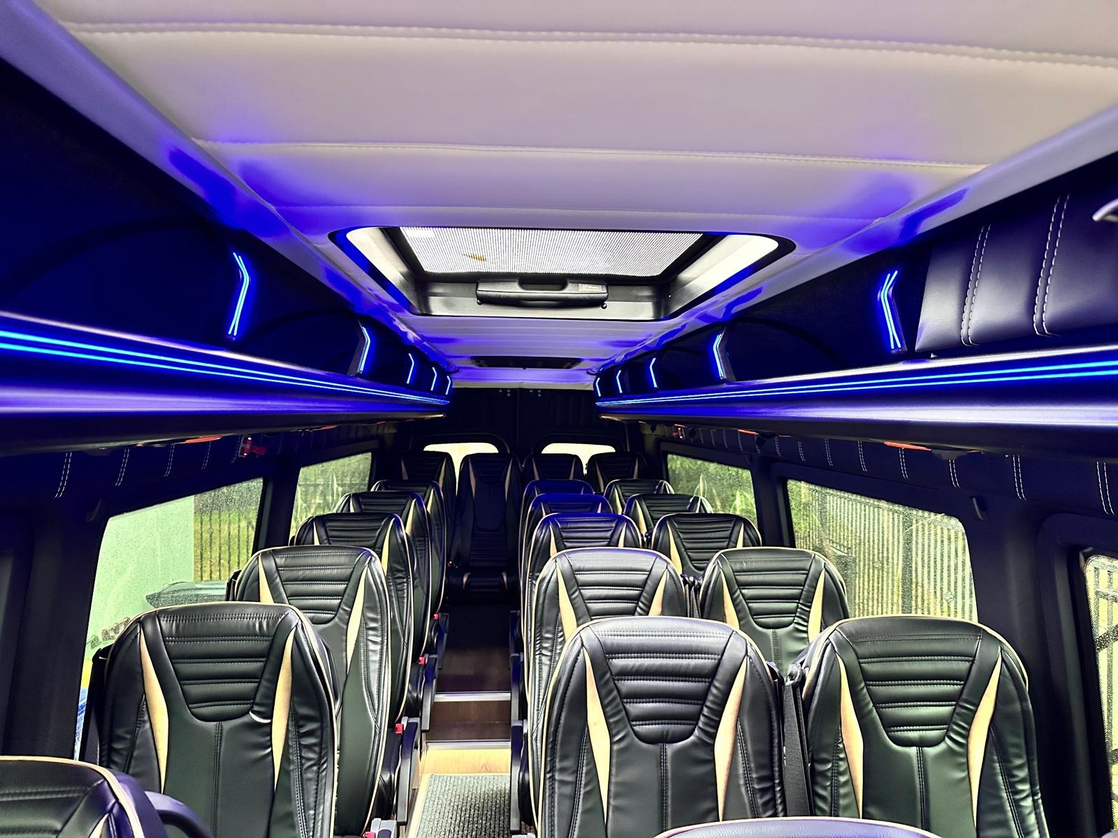 Wnętrze luksusowego busa z czarnymi skórzanymi fotelami z beżowymi wstawkami i nastrojowym, niebieskim oświetleniem LED. Widok na okna i sufit z oknem dachowym.