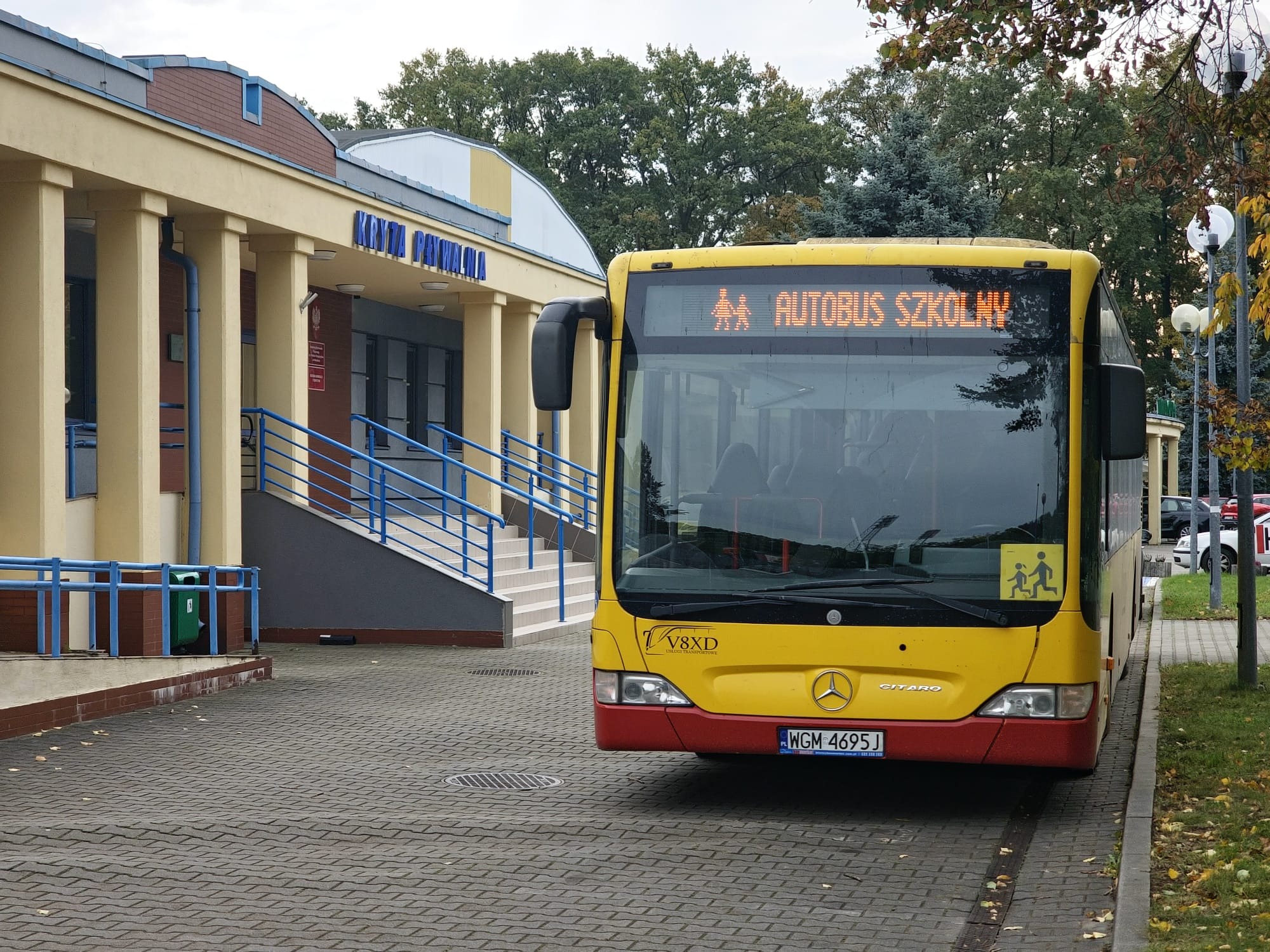 Żółty autobus szkolny Mercedes-Benz Citato z napisem LED i piktogramami dzieci, zaparkowany przed budynkiem z napisem 'Kryta Pływalnia'.