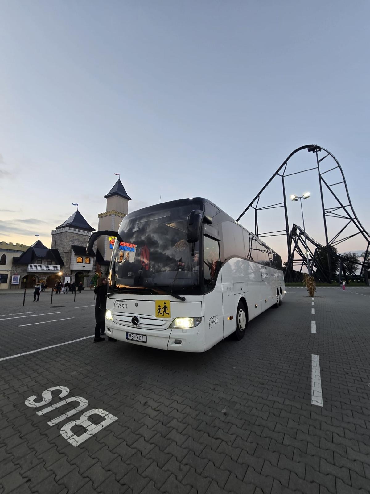 Biały autokar Mercedes-Benz V8XD na parkingu z napisem 'BUS', w tle park rozrywki z kolejką górską i zamkiem. Tabliczka z symbolem dzieci.