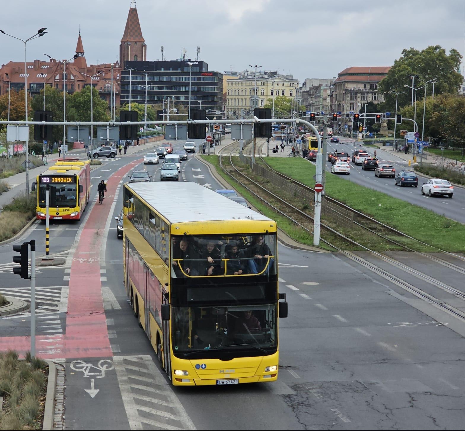 Żółty, dwupiętrowy autobus MAN na tle miejskiego krajobrazu z czerwonym pasem dla rowerów i tramwajowymi torami. Widoczni pasażerowie na górnym pokładzie.