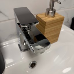 Dom Bez Plamy - Błyszcząca, chromowana bateria umywalkowa o kwadratowym kształcie, obok dozownik mydła w drewnianej obudowie na tle białej umywalki i marmurowej ściany.