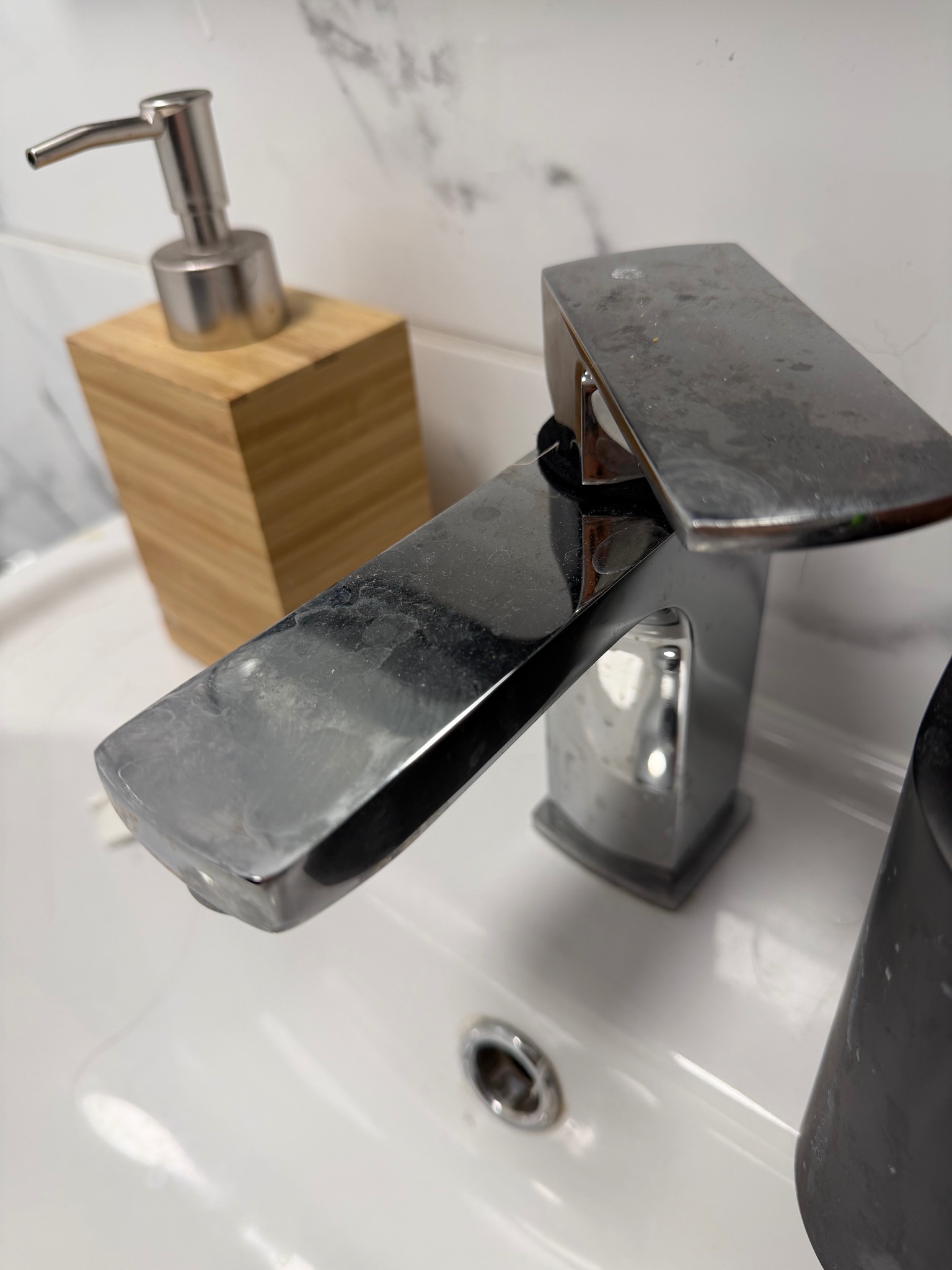 Zacieki i osad na chromowanej baterii umywalkowej, obok dozownik mydła w drewnianej obudowie. Widoczne ślady użytkowania na białej umywalce i marmurowym tle.