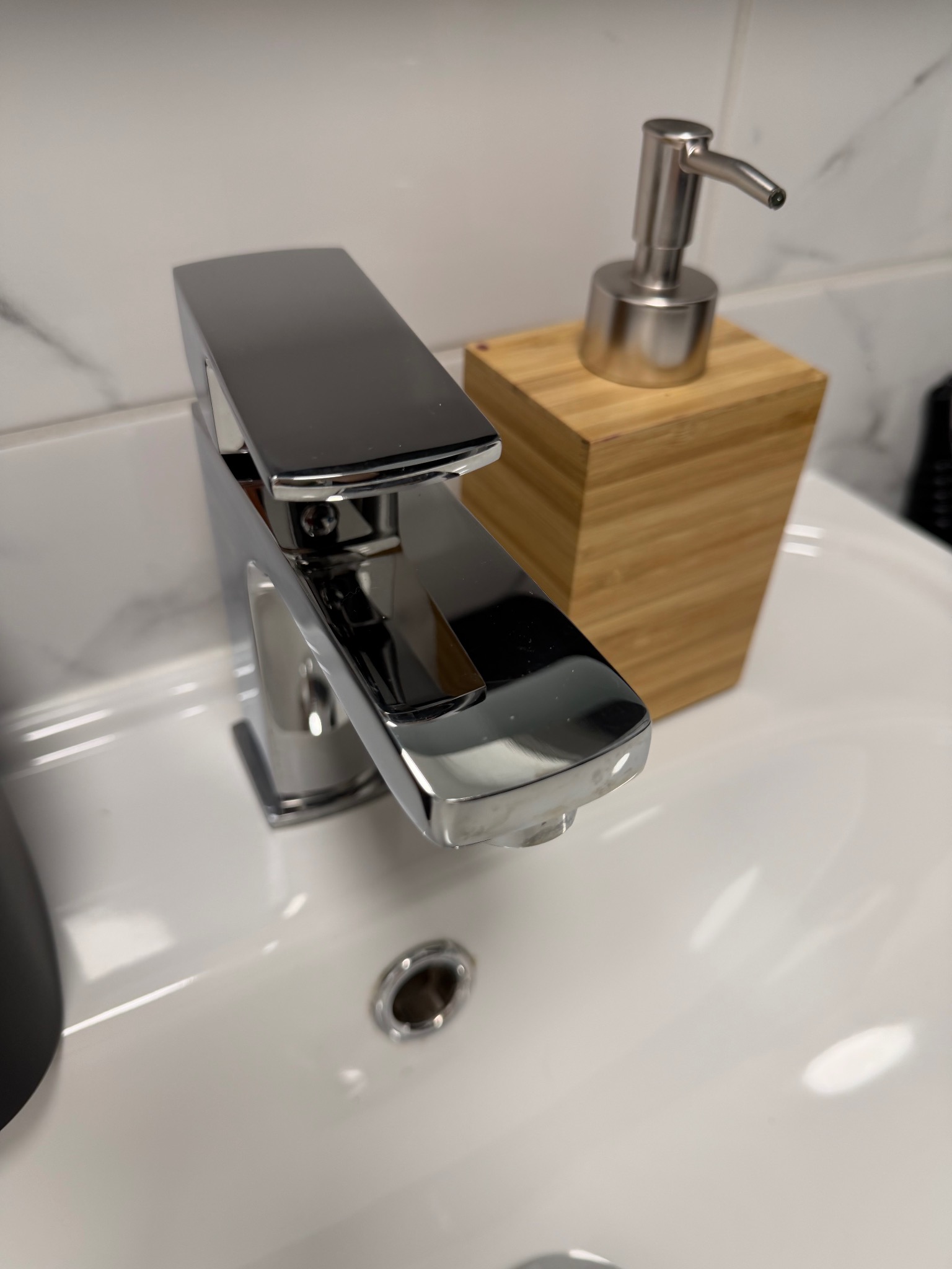 Błyszcząca, chromowana bateria umywalkowa o kwadratowym kształcie, obok dozownik mydła w drewnianej obudowie na tle białej umywalki i marmurowej ściany.