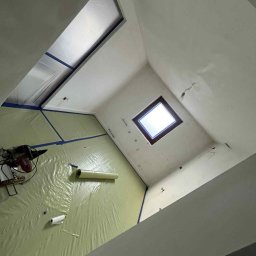 Dbw home - Przygotowanie do malowania poddasza: folia ochronna na podłodze, okna dachowe, świeżo pomalowane ściany. Widok z dołu, ukośne ujęcie.