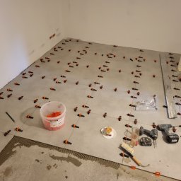 Dbw home - Układanie szarych płytek podłogowych z użyciem systemu poziomowania. Widoczne narzędzia, wiadro z klejem, poziomica i przekąska na talerzyku. Częściowo ułożona podłoga.