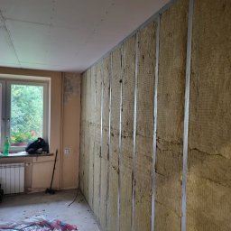 Dbw home - Ocieplenie ściany wełną mineralną, widoczne metalowe profile. Na podłodze folia ochronna, w tle okno z widokiem na zieleń. Etap prac wykończeniowych wewnątrz pomieszczenia.