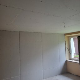 Dbw home - Pokój w trakcie prac wykończeniowych: ściany i sufit pokryte płytami gipsowo-kartonowymi, widoczne łączenia i otwory po wkrętach. Kabel zwisa z sufitu, okno z parapetem.