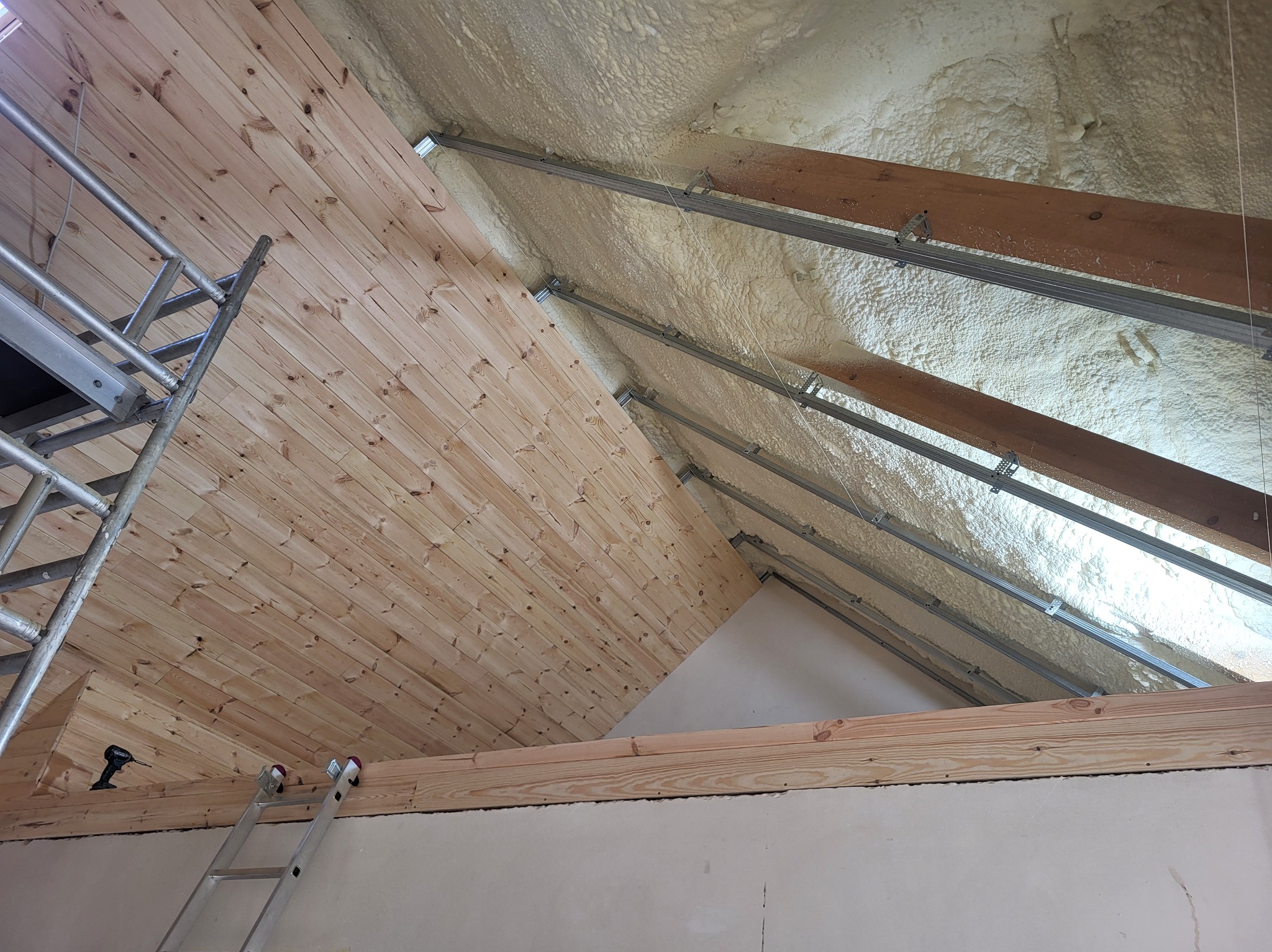 Drewniana boazeria na poddaszu z widoczną izolacją piankową, metalowymi profilami i drabiną. Ujęcie z dołu, ukazujące etap prac wykończeniowych.