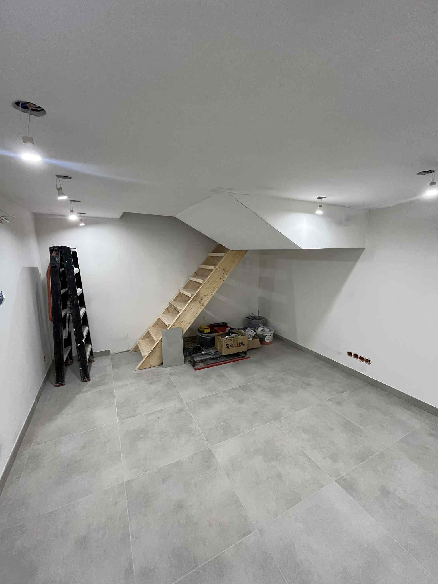 Wykończone wnętrze z szarą podłogą, drewnianymi schodami i świeżo pomalowanymi ścianami. Widoczna drabina i materiały budowlane. Oświetlenie sufitowe.