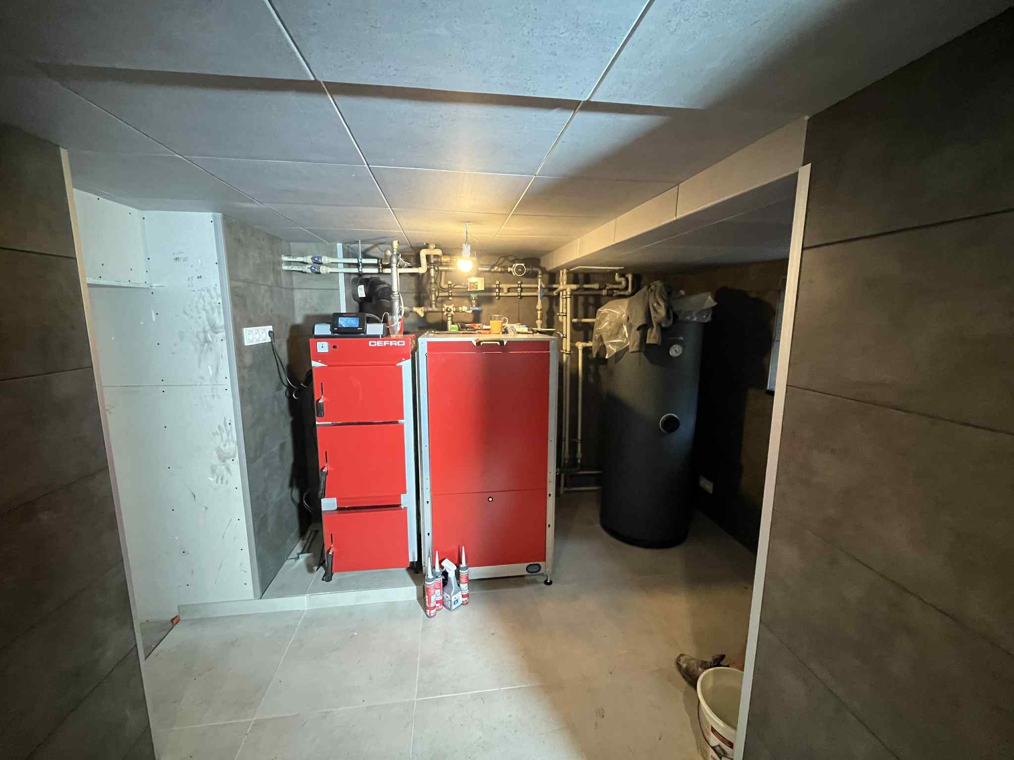 Czerwony piec CO marki Defro w kotłowni z szarymi płytkami na ścianach i podłodze. Widoczne rury instalacji grzewczej i zbiornik akumulacyjny.