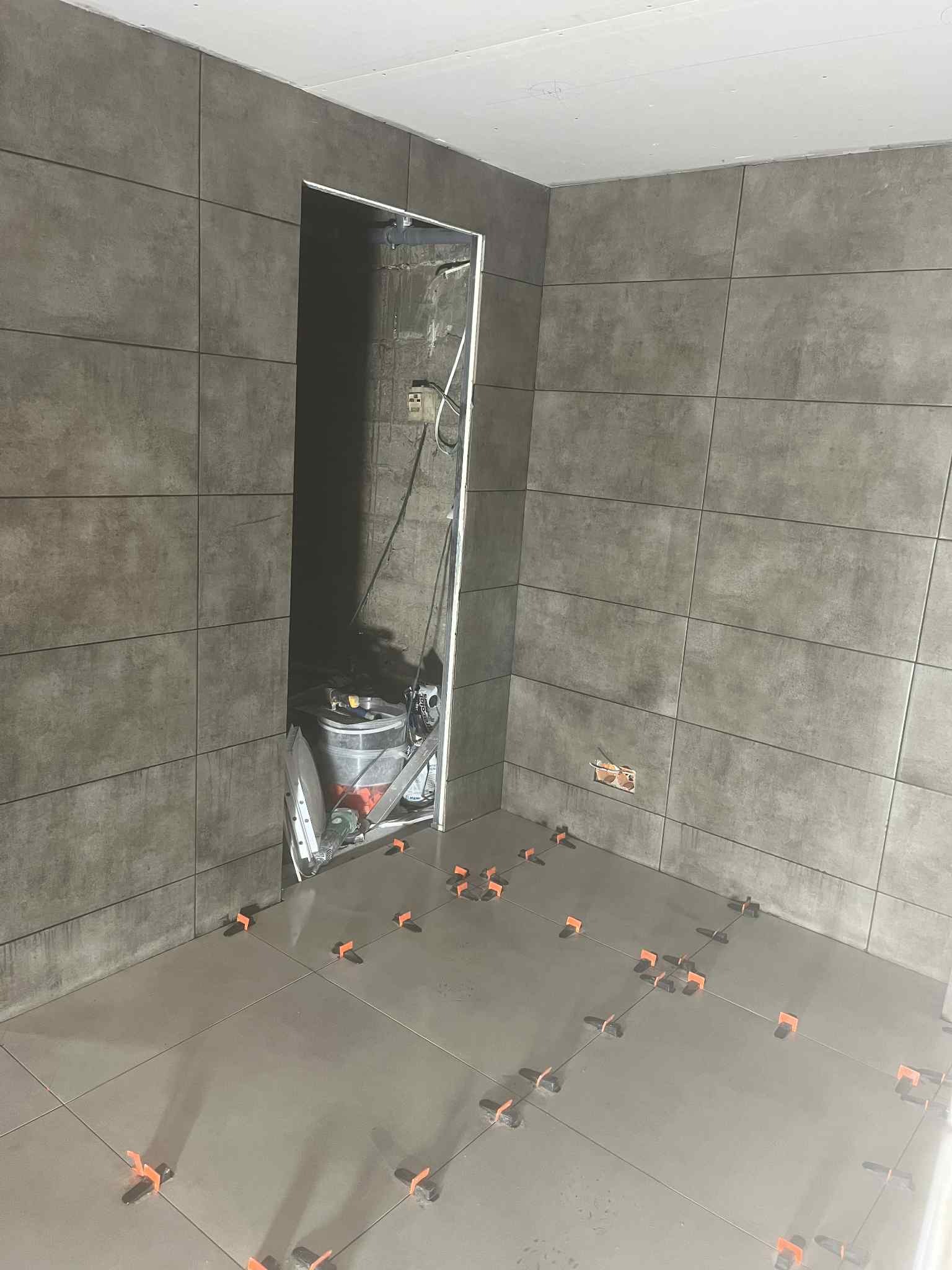 Układanie szarych płytek w łazience z użyciem klipsów poziomujących. Widoczne narzędzia i wiadro w otworze drzwiowym. Ściany również wyłożone płytkami.