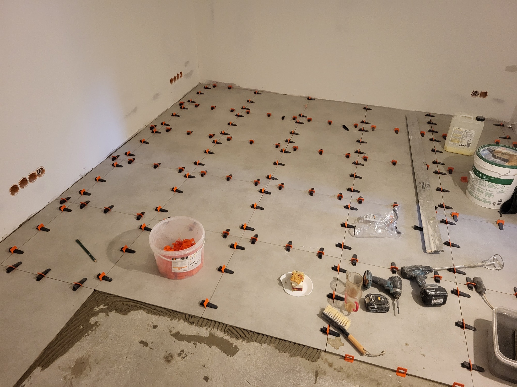 Układanie szarych płytek podłogowych z użyciem systemu poziomowania. Widoczne narzędzia, wiadro z klejem, poziomica i przekąska na talerzyku. Częściowo ułożona podłoga.