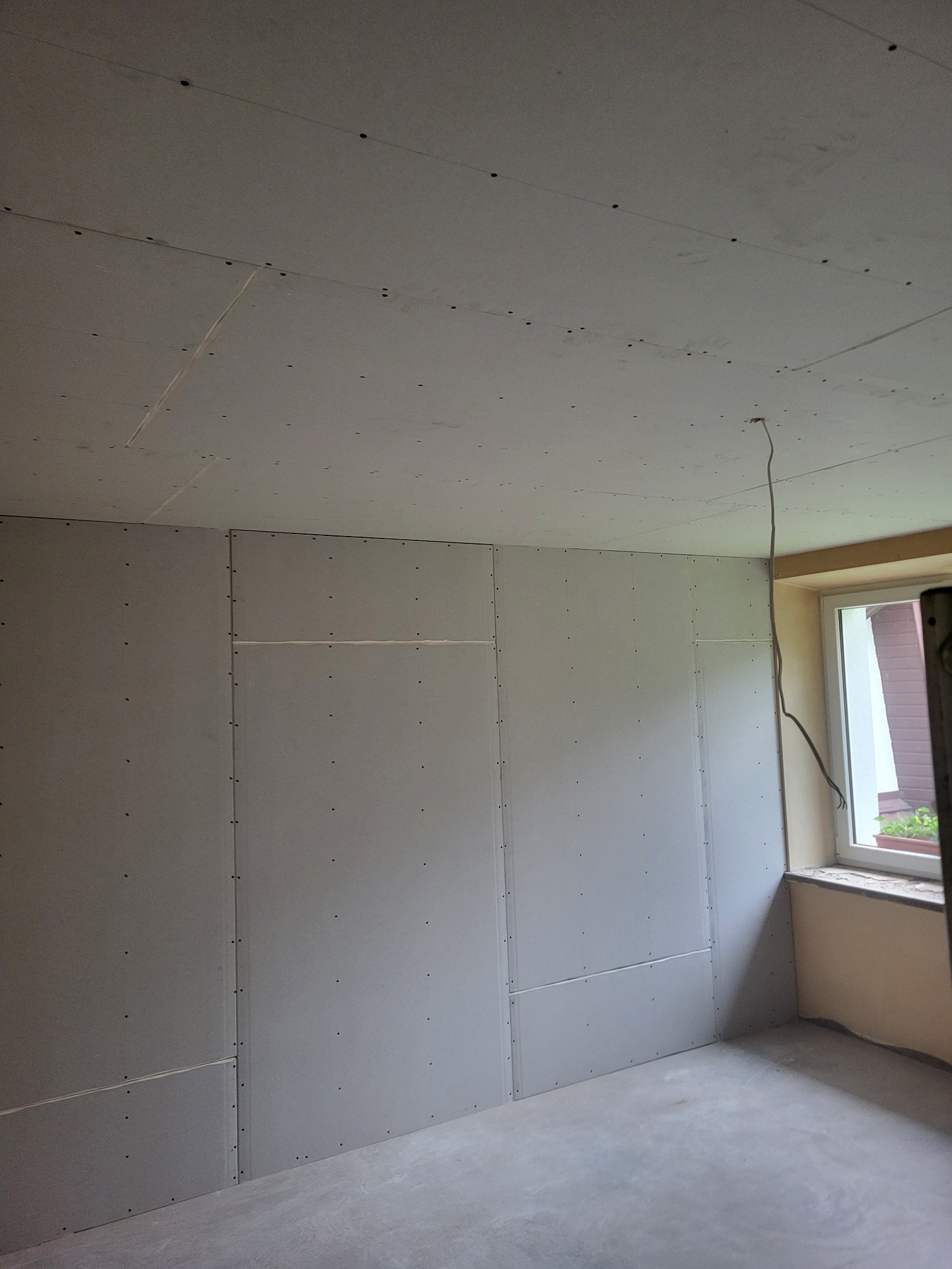 Pokój w trakcie prac wykończeniowych: ściany i sufit pokryte płytami gipsowo-kartonowymi, widoczne łączenia i otwory po wkrętach. Kabel zwisa z sufitu, okno z parapetem.