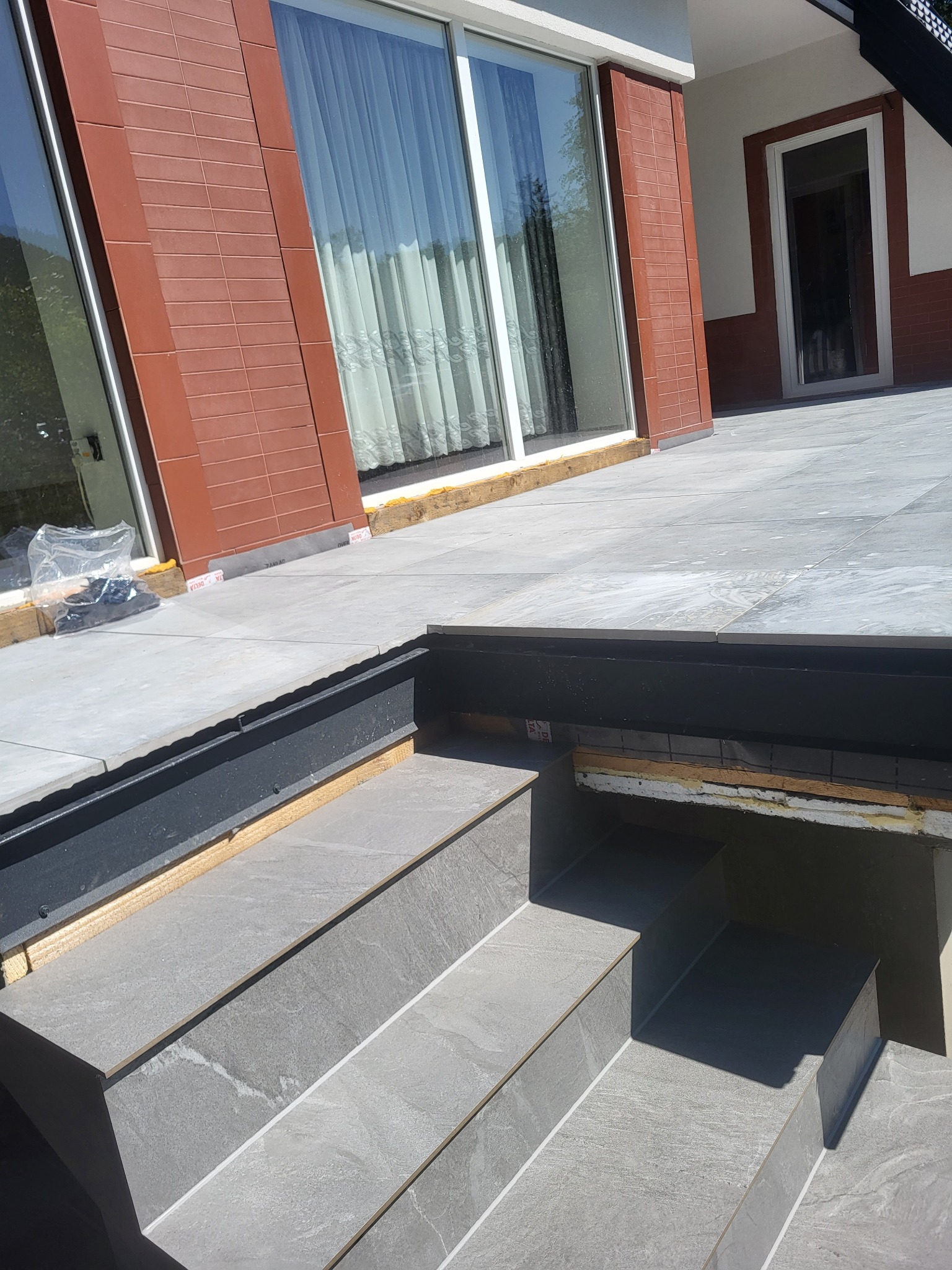 Wykończony taras z szarych płytek ceramicznych, schody z płytek, widoczne detale wykończeniowe, okno tarasowe z zasłonami, elewacja z cegły klinkierowej.