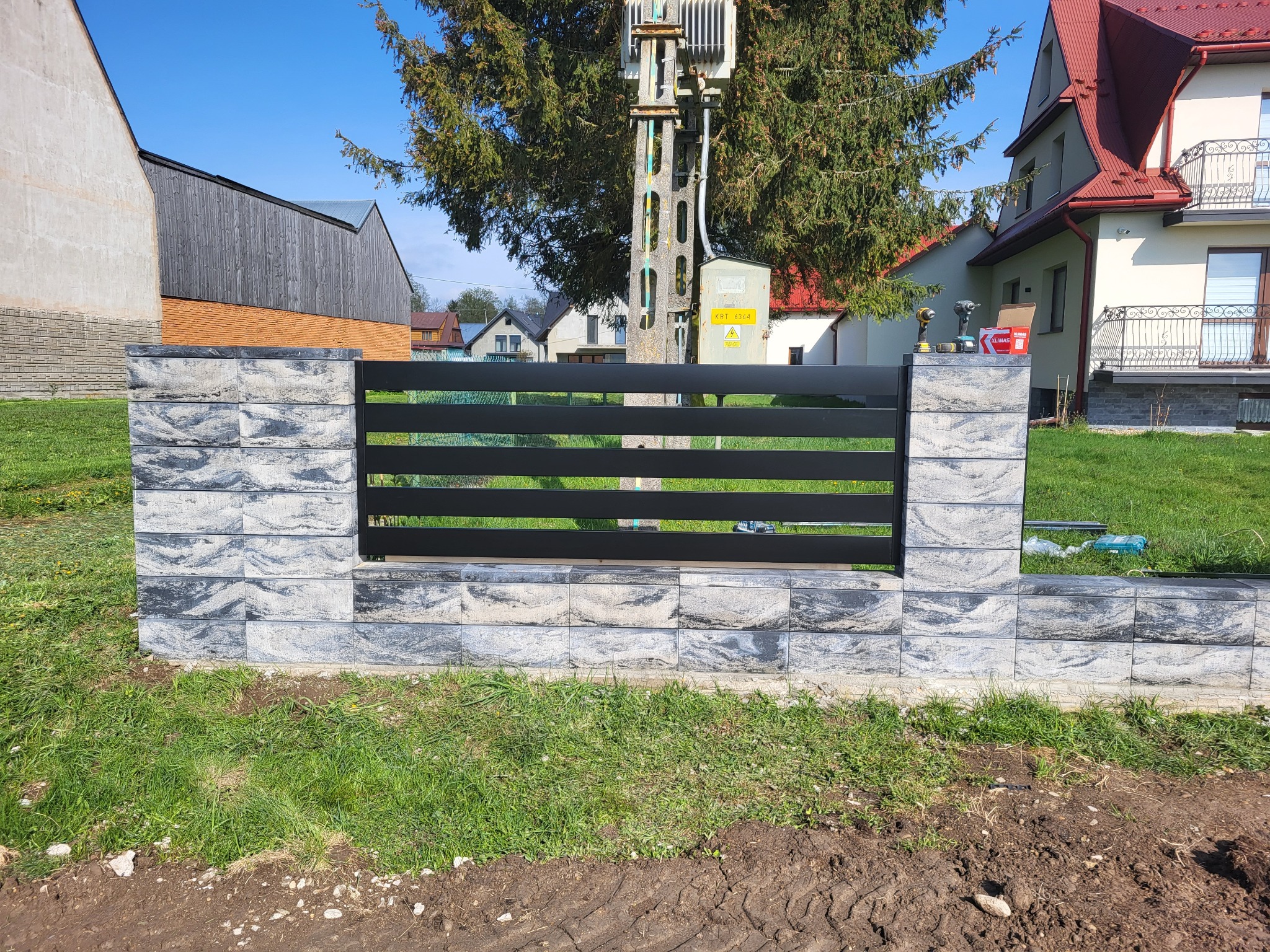 Nowoczesne ogrodzenie z szarej kostki i czarnych, poziomych paneli, z narzędziami na słupku. Trawa i budynki w tle. Słoneczny dzień.