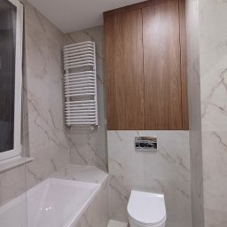 Łukasz Machnio - Wykończona łazienka z wanną, grzejnikiem drabinkowym, toaletą i szafką wiszącą. Płytki imitujące marmur na ścianach i podłodze. Nowoczesny design.