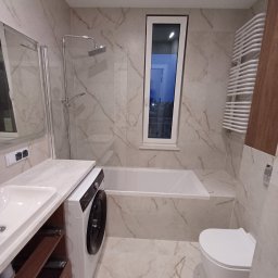 Łukasz Machnio - Wykończona łazienka w jasnych marmurkowych płytkach z wanną, toaletą, umywalką z szafką i pralką. Wąskie okno w głębi. Grzejnik drabinkowy po prawej.