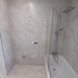 Łukasz Machnio - Wykończona łazienka z wanną i szklanym parawanem, wyłożona płytkami imitującymi marmur. Widoczne baterie, deszczownica i przełączniki. Minimalistyczny design.