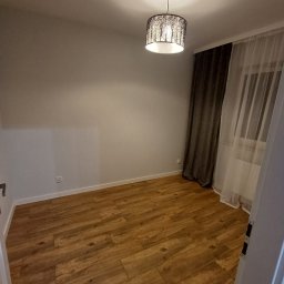 Łukasz Machnio - Jasne drewniane panele podłogowe w pokoju z szarą ścianą, oświetlone lampą z motywem drzew. Okno z zasłonami w tle. Widok zza otwartych drzwi.