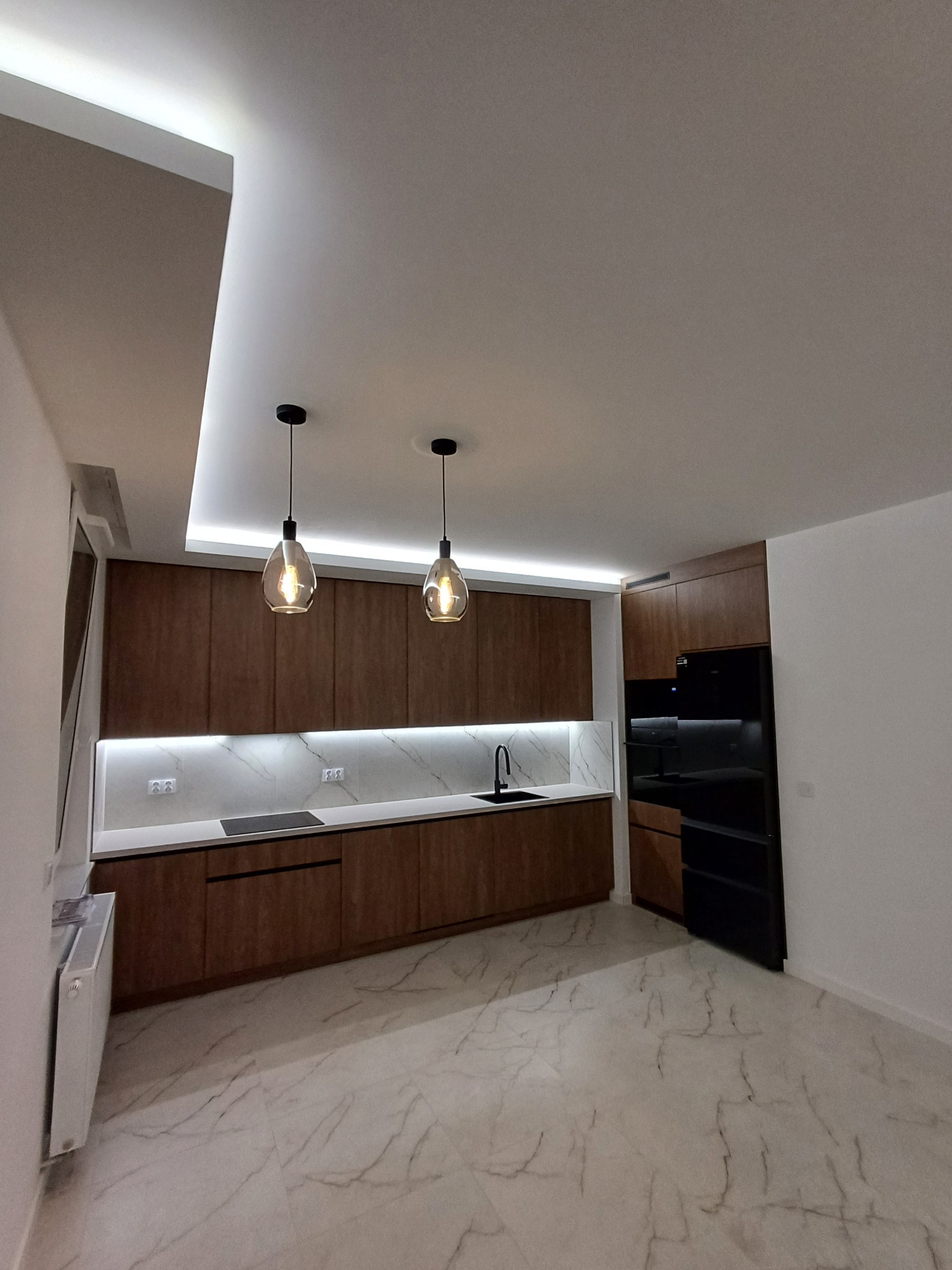 Nowoczesna kuchnia z drewnianymi szafkami, marmurowym blatem i podświetleniem LED. Dwie wiszące lampy z żarówkami Edisona. Czarny sprzęt AGD.