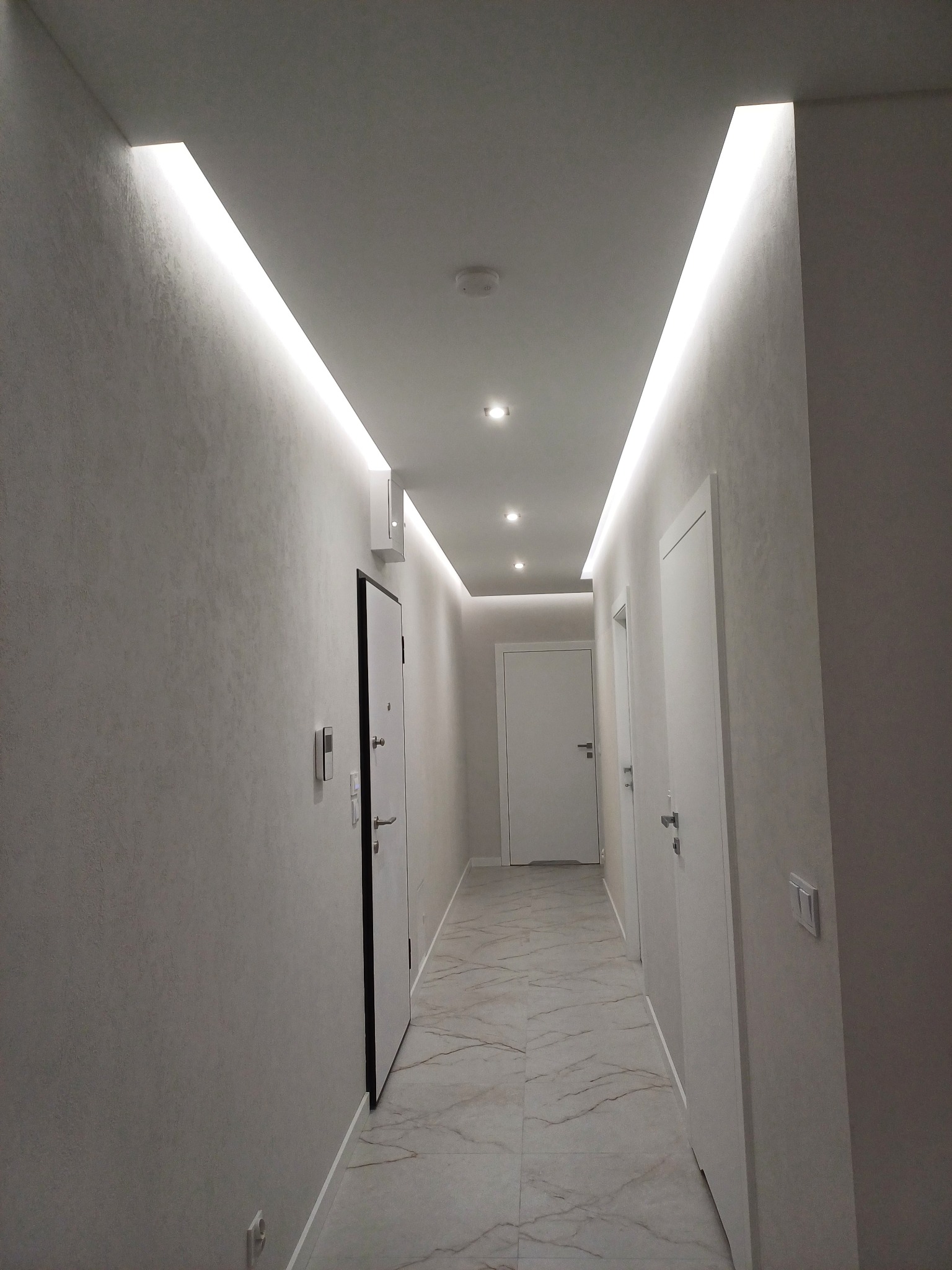 Nowoczesny korytarz z oświetleniem LED w suficie, marmurową podłogą i białymi drzwiami. Ściany w odcieniach szarości z delikatną fakturą. Minimalistyczny design.