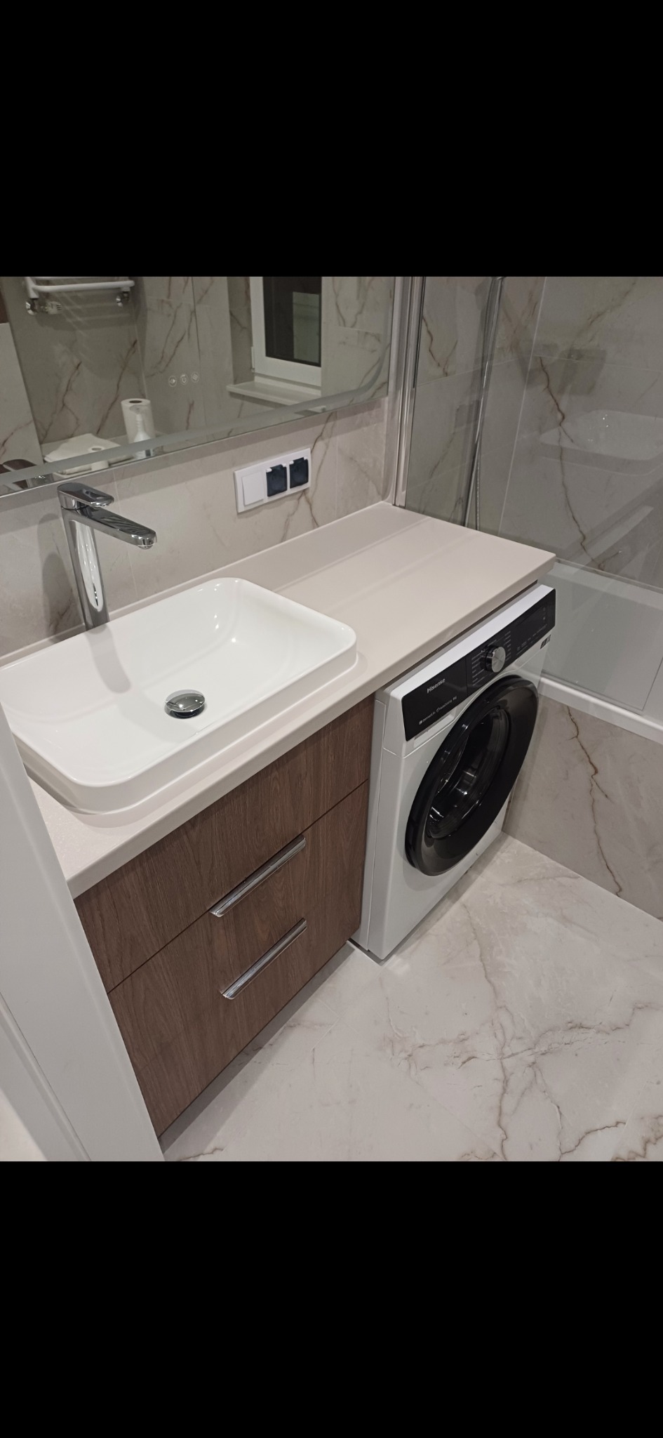 Nowoczesna łazienka z umywalką nablatową, szafką z drewnopodobnym frontem i pralką pod blatem. Marmurowe płytki na ścianach i podłodze.