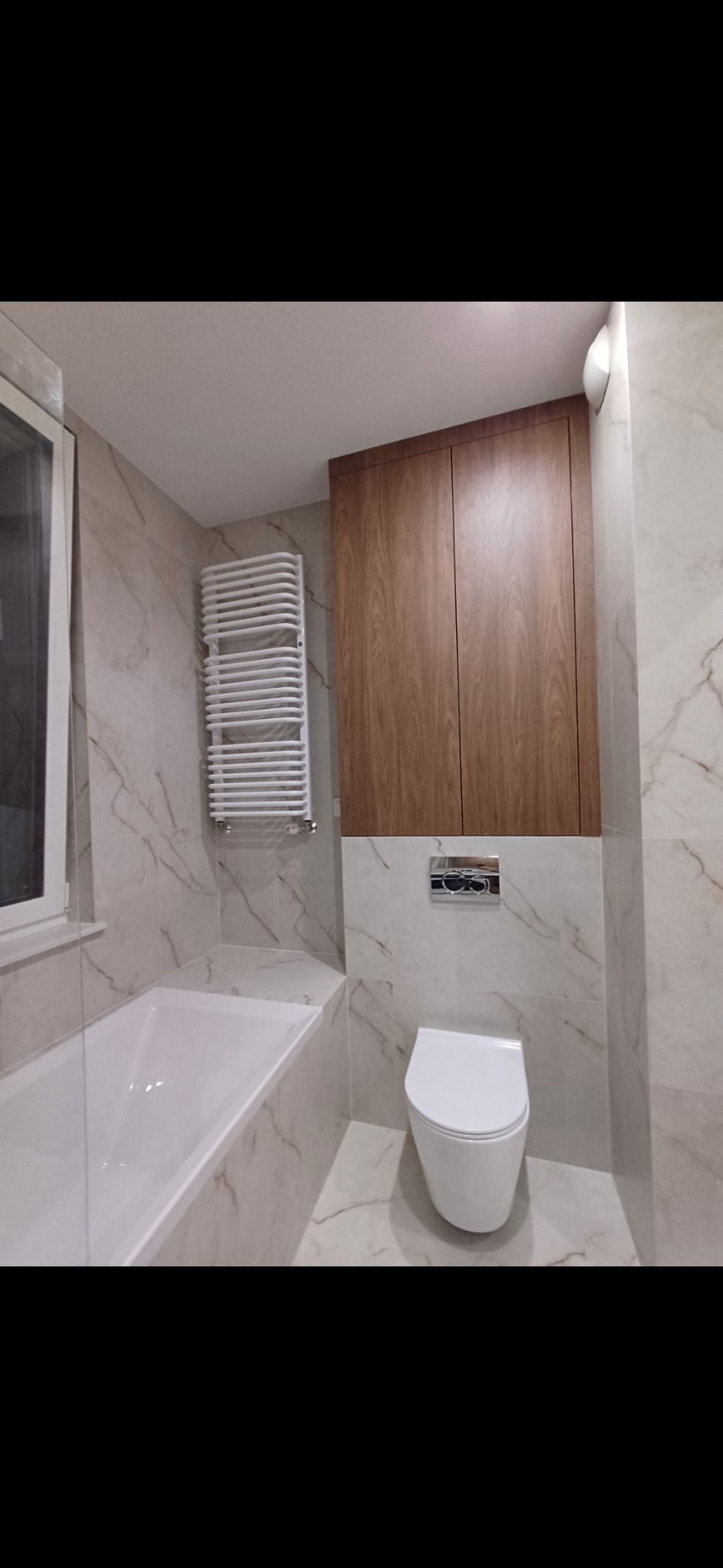 Wykończona łazienka z wanną, grzejnikiem drabinkowym, toaletą i szafką wiszącą. Płytki imitujące marmur na ścianach i podłodze. Nowoczesny design.