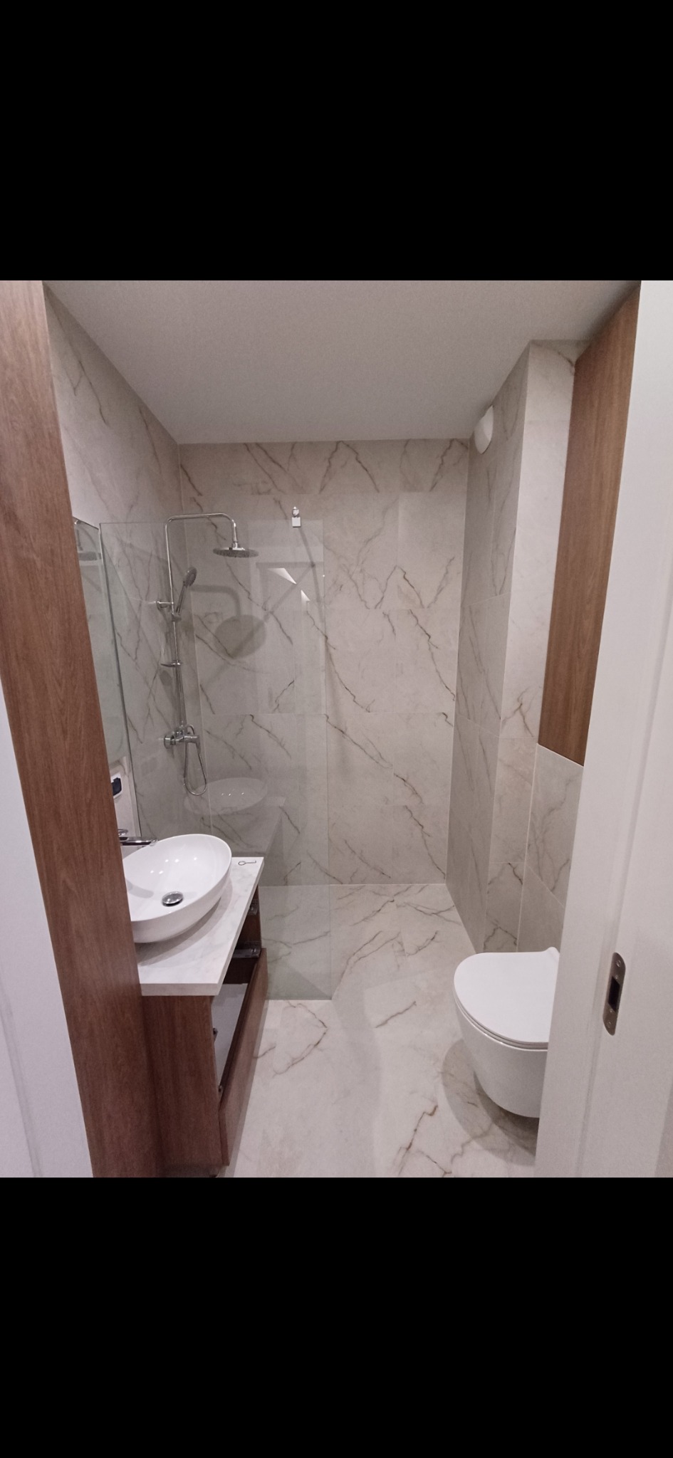 Wykończona łazienka z kabiną prysznicową typu walk-in, umywalką nablatową i toaletą. Marmurowe płytki na ścianach i podłodze. Elementy wykończenia w kolorze drewna.