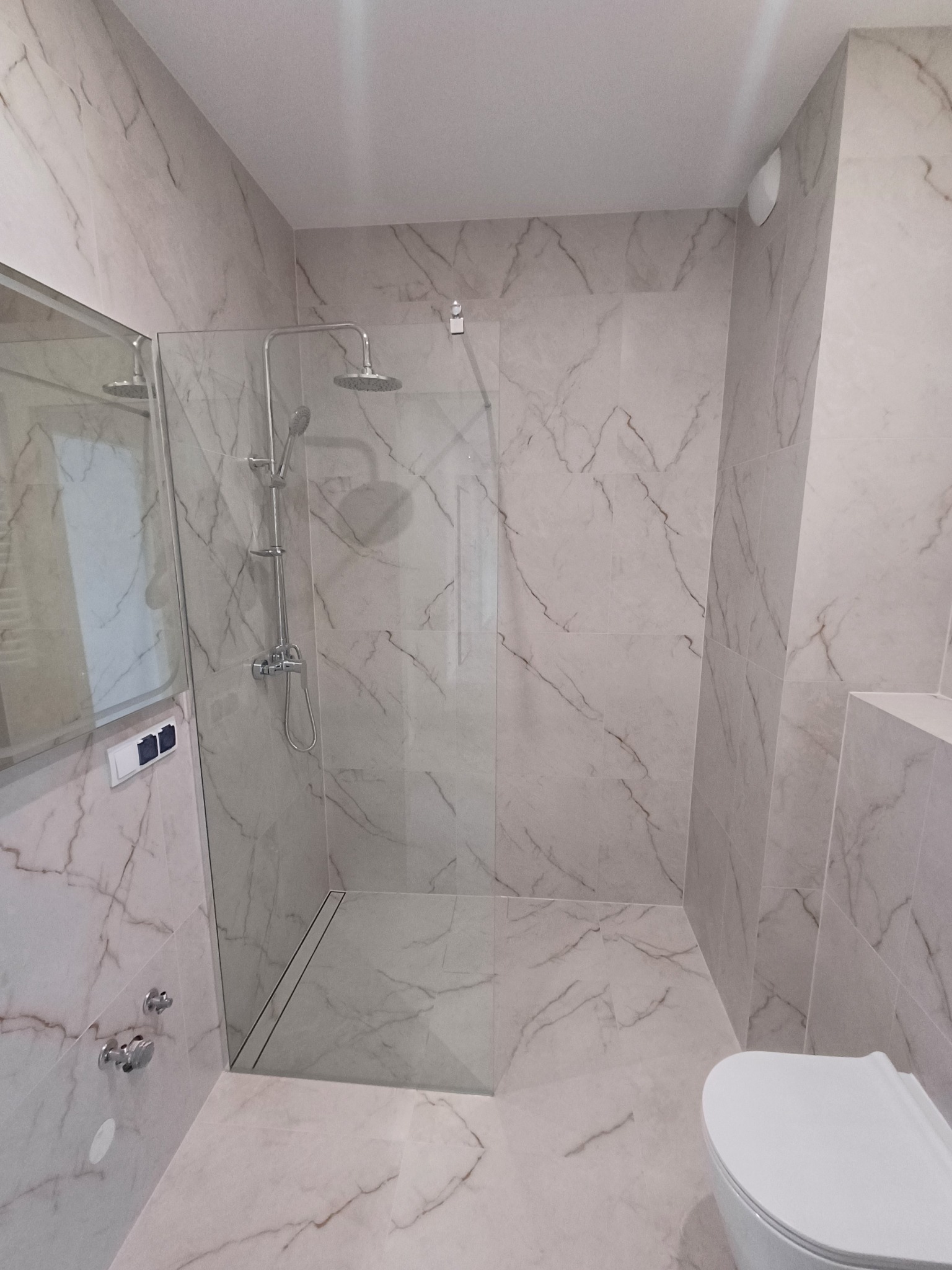 Wykończona łazienka z kabiną typu walk-in, wyłożona płytkami imitującymi marmur. Widoczna armatura, toaleta i szklana ścianka kabiny.