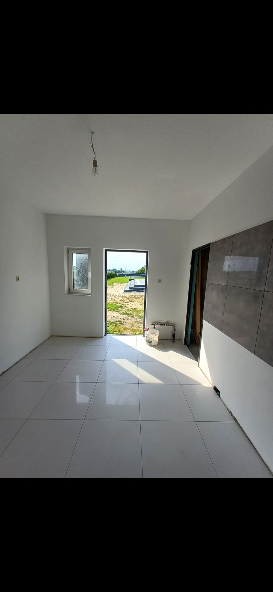 Jasne wnętrze z białymi płytkami podłogowymi i częściowo wykończoną ścianą z szarymi płytkami. Widoczne okno i otwarte drzwi z widokiem na zewnątrz.