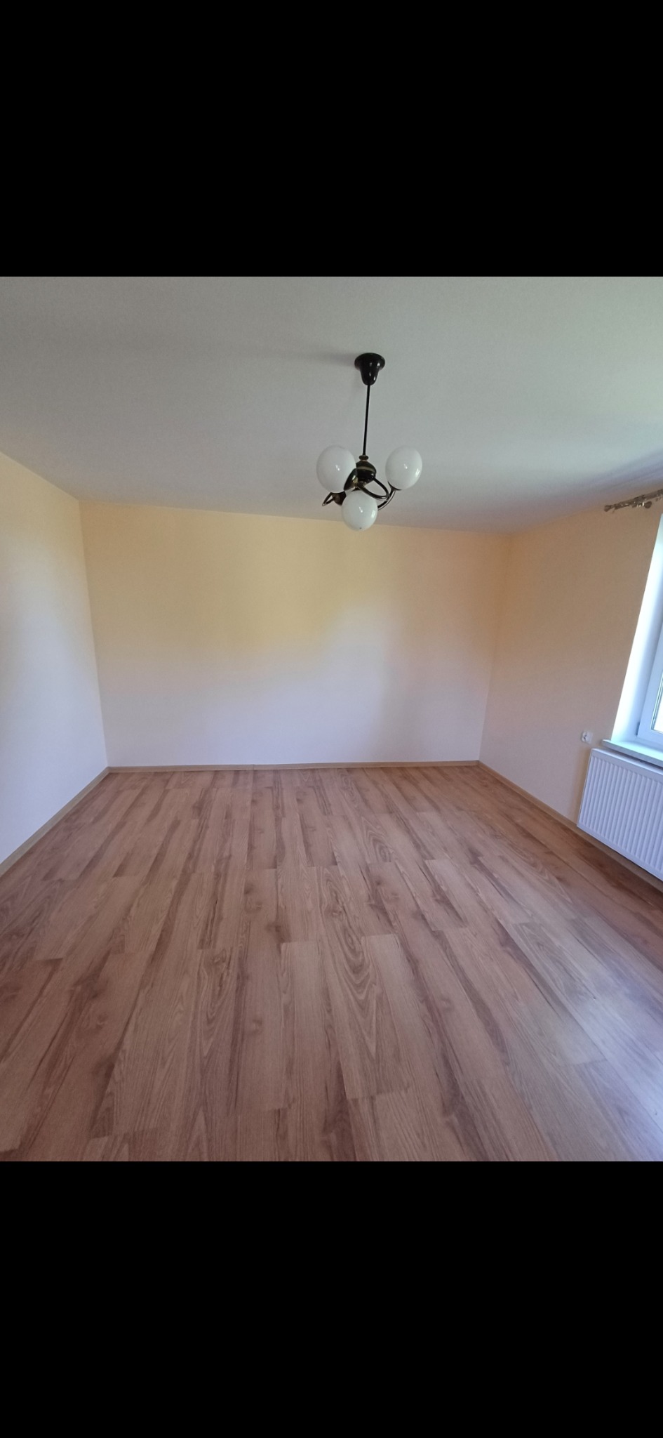 Jasne wnętrze po remoncie: parkiet laminowany w odcieniach brązu, kremowe ściany i czarna lampa sufitowa z białymi kloszami. Widok na grzejnik pod oknem.