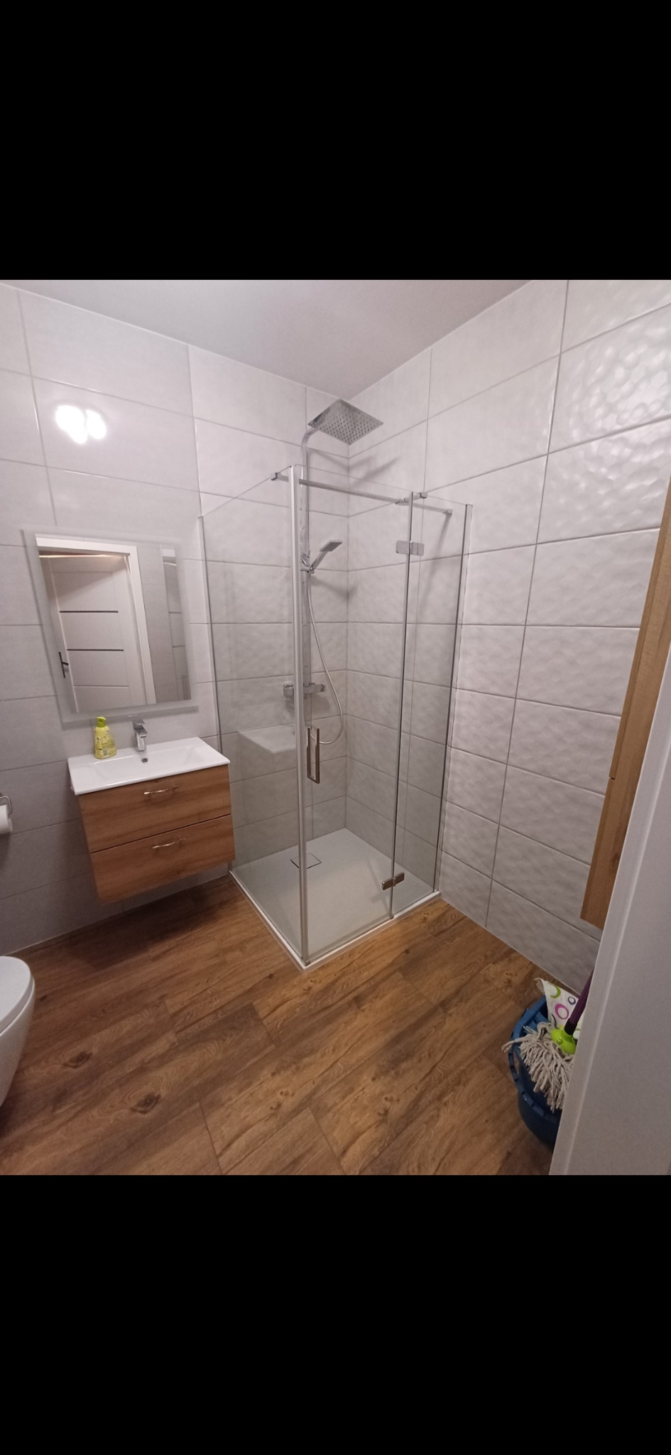 Nowoczesna łazienka po remoncie: kabina prysznicowa ze szklanymi ścianami, drewnopodobna podłoga, umywalka z szafką i lustro. Białe płytki na ścianach.