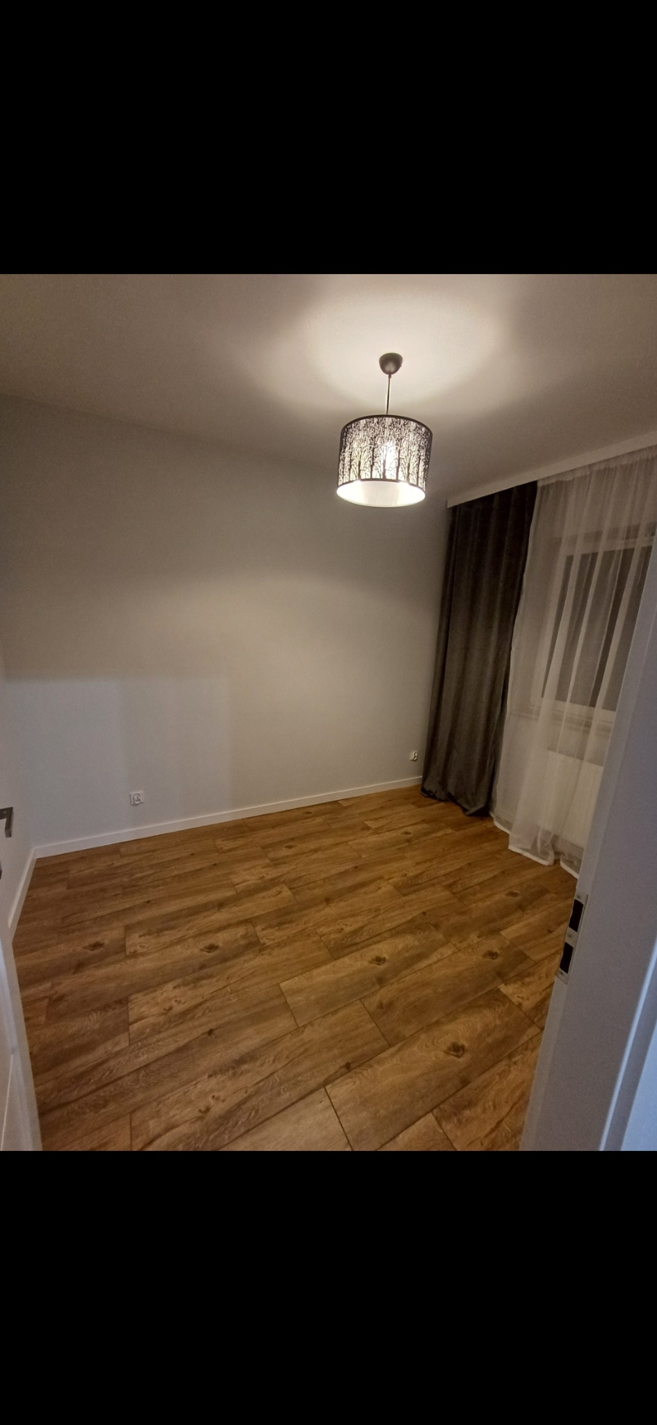 Jasne drewniane panele podłogowe w pokoju z szarą ścianą, oświetlone lampą z motywem drzew. Okno z zasłonami w tle. Widok zza otwartych drzwi.