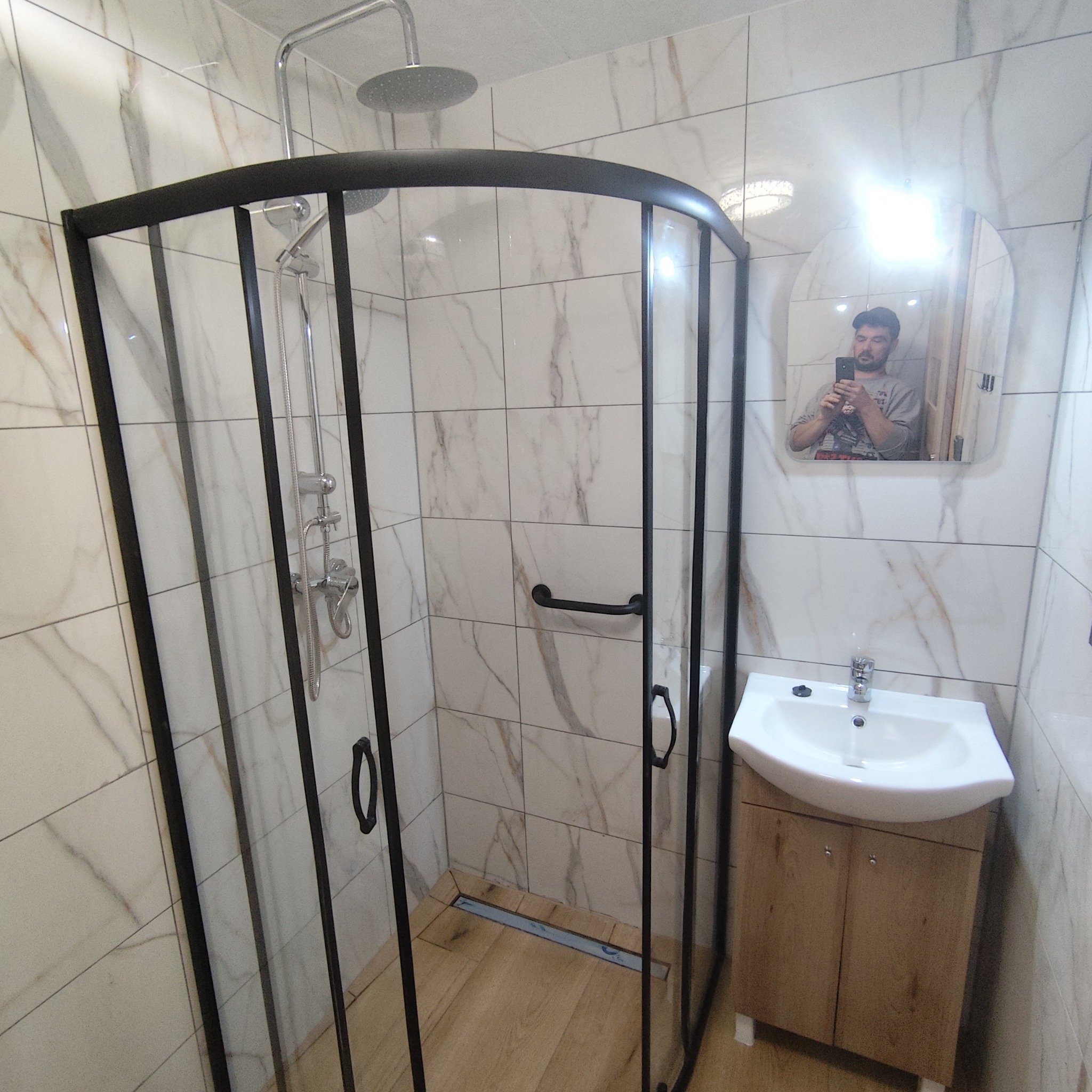 Nowoczesna kabina prysznicowa z czarnymi ramami i deszczownicą, umywalka z szafką w wykończonej łazience z marmurowymi płytkami. W lustrze odbicie osoby robiącej zdjęcie.