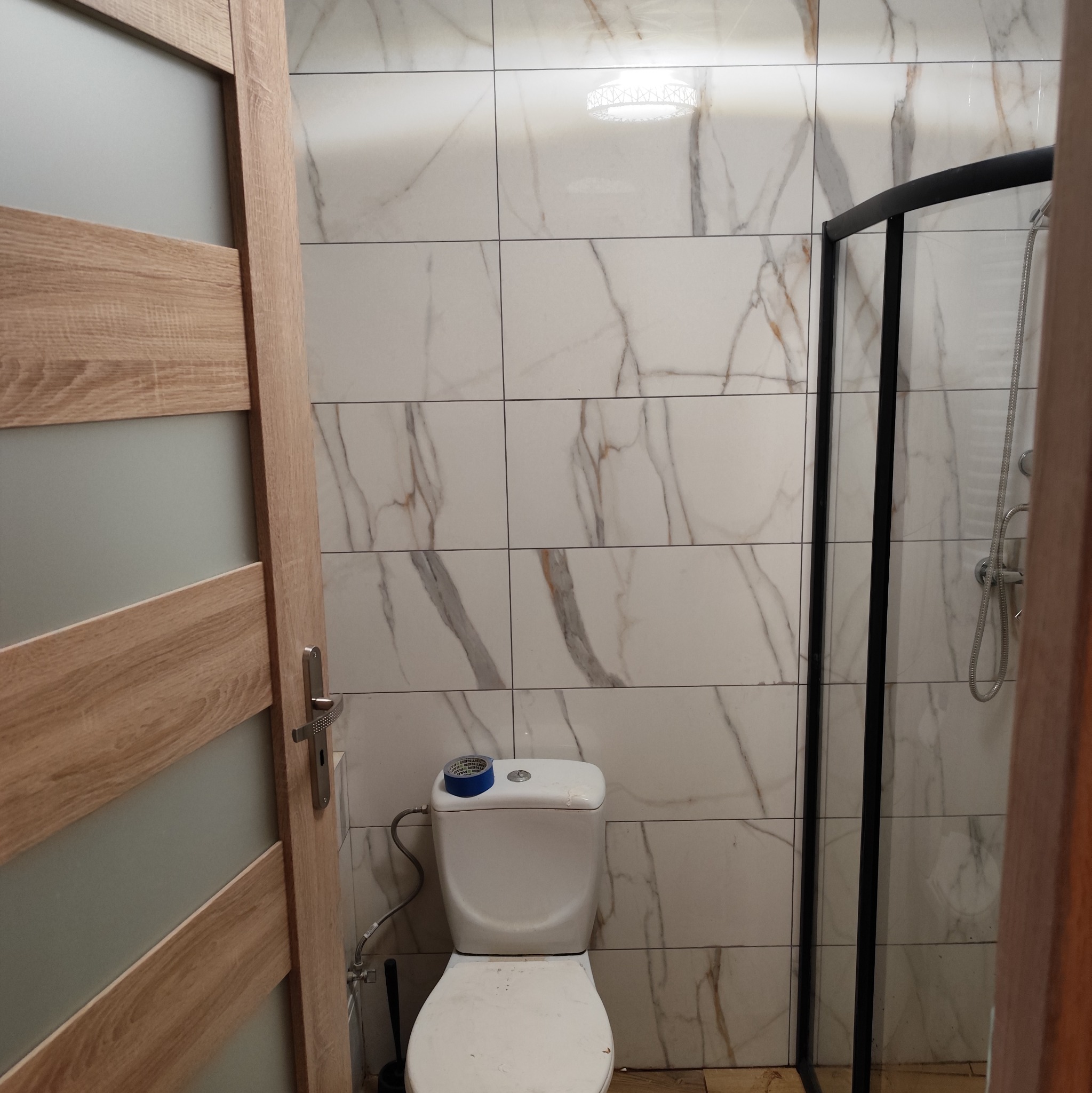 Wykończona łazienka z marmurowymi płytkami, kabiną prysznicową z czarnymi ramami i toaletą z niebieską taśmą. Widok zza drewnianych drzwi.