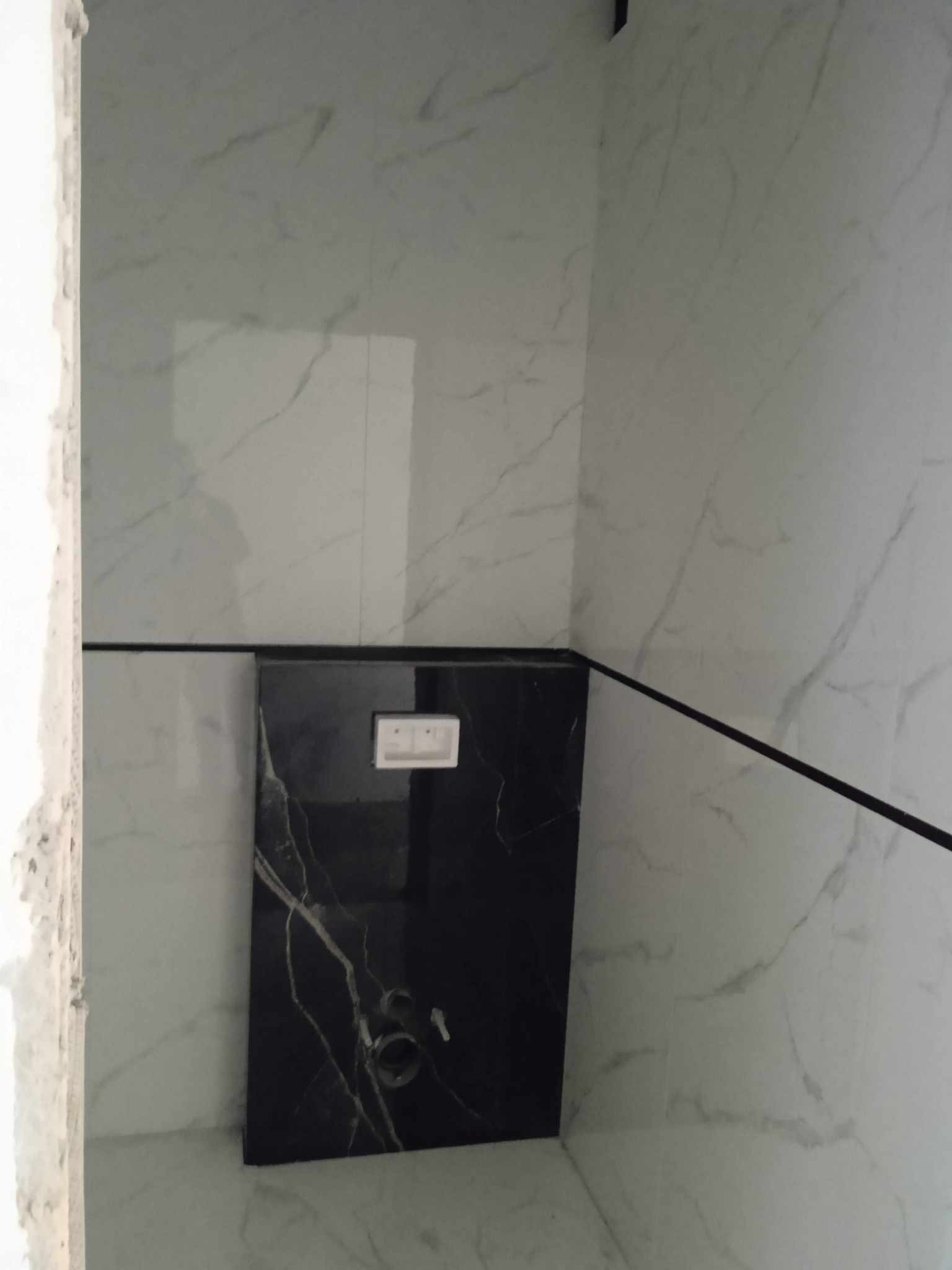 Wykończona ściana w łazience z białych płytek imitujących marmur i czarnym panelem z marmurowym wzorem, z widocznymi przyłączami i włącznikiem.